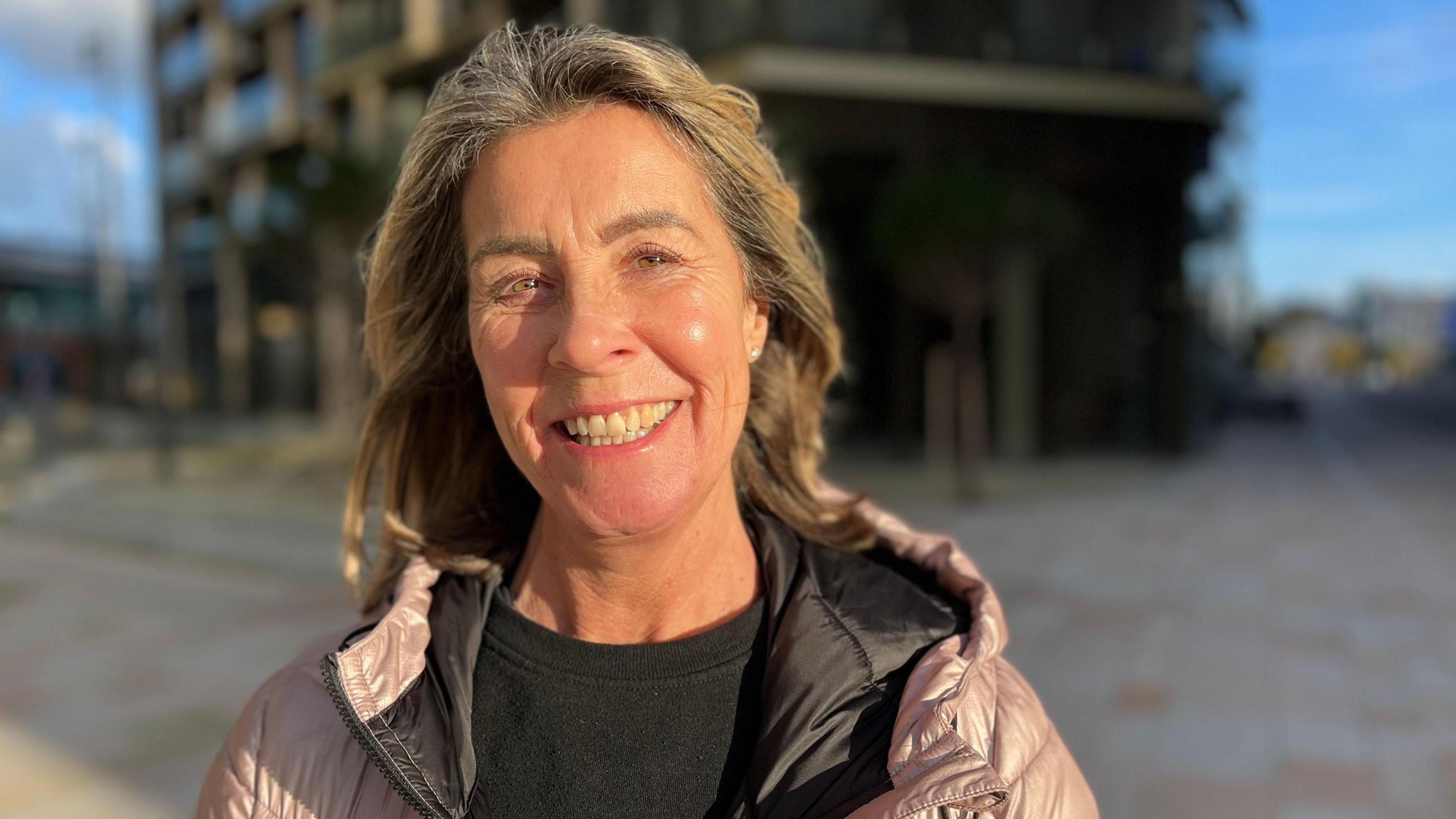 Ruth Young is a woman with brown, shoulder-length hair. She is smiling into the camera. She is wearing a black t shirt and a purple puffer coat. The background is an out-of-focus building at the waterfront with a paved path around it.