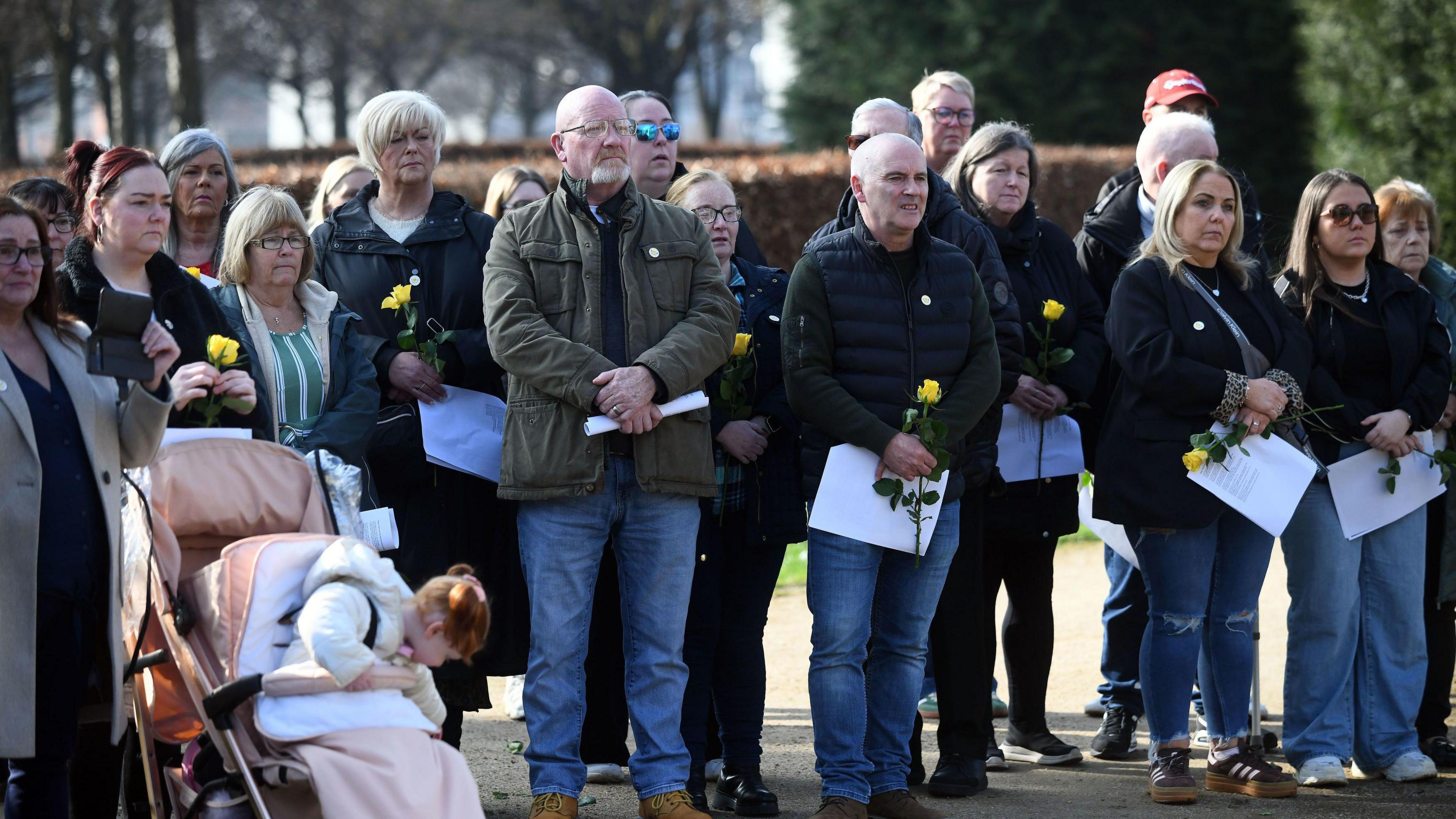 A group of people carrying yellow roses and papers, looking solem, with a child in a buggy on the left of shot