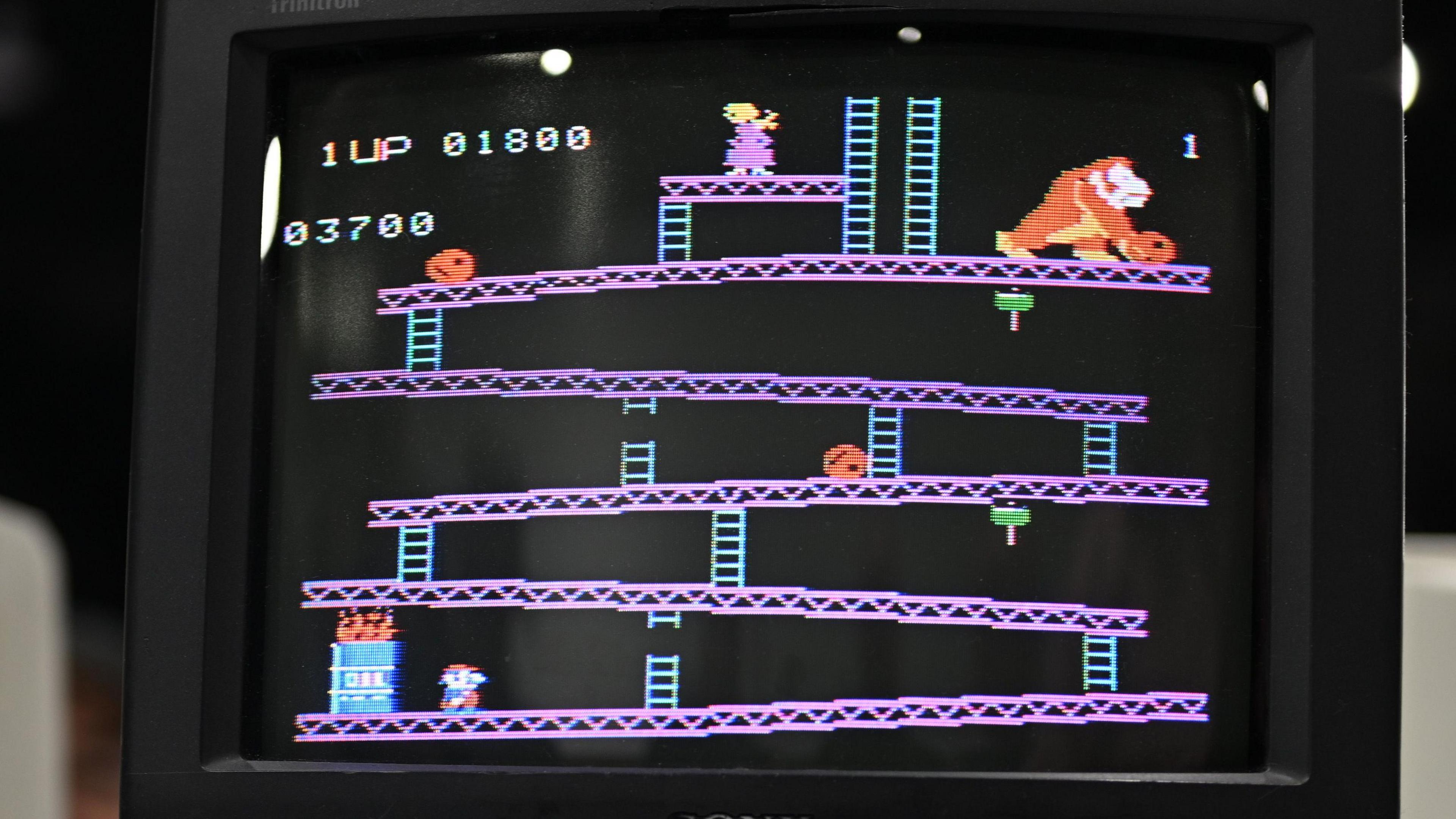 A photo of a screen showing the arcade game Donkey Kong, with a small pixelated character wearing overalls running up scaffolding and ladders, and characters of a woman and giant gorilla at the top