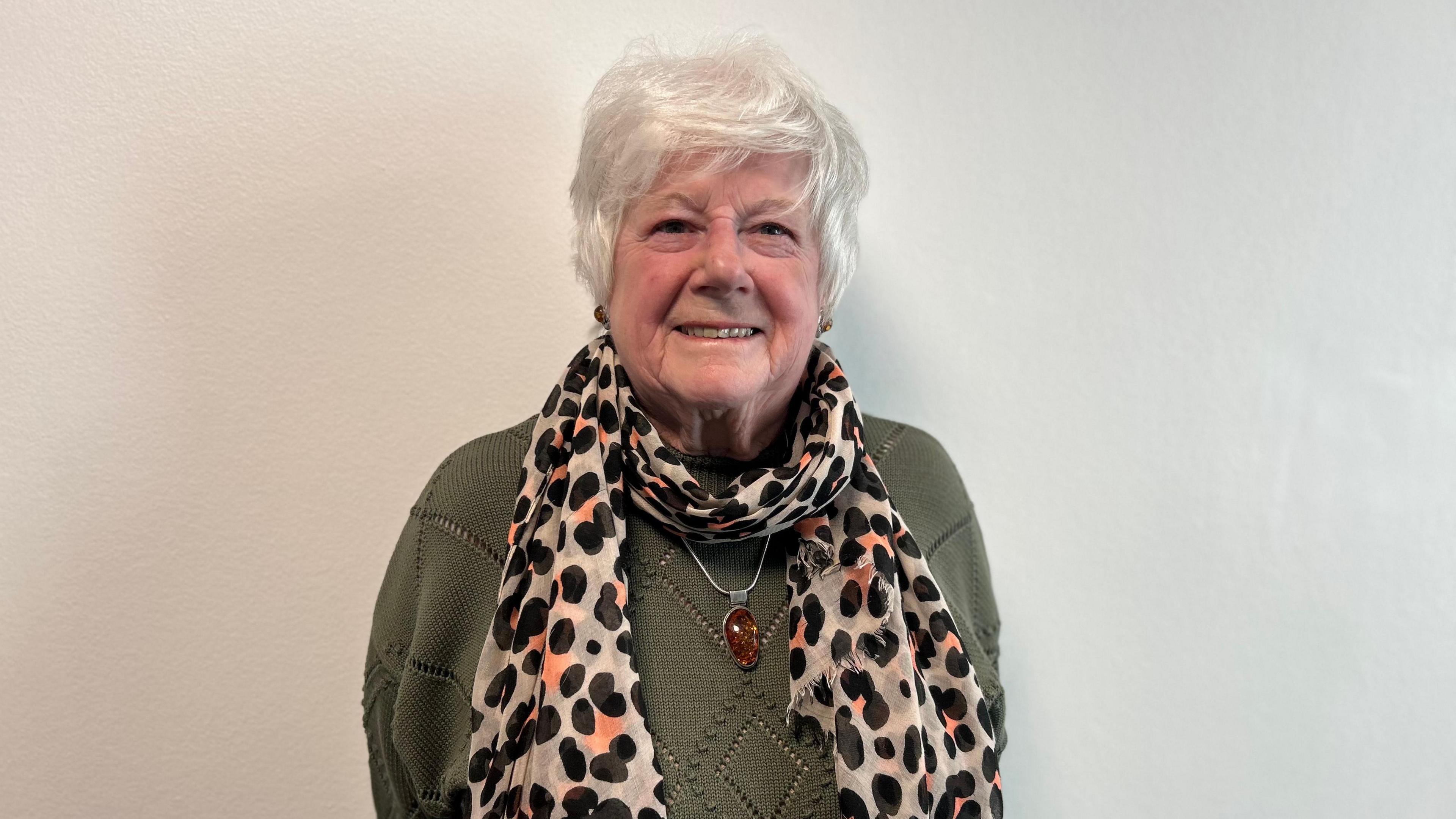 A woman with grey hair and wearing a green jumper, a spotty scarf and a large amber necklace, smiling at the camera against a white background.