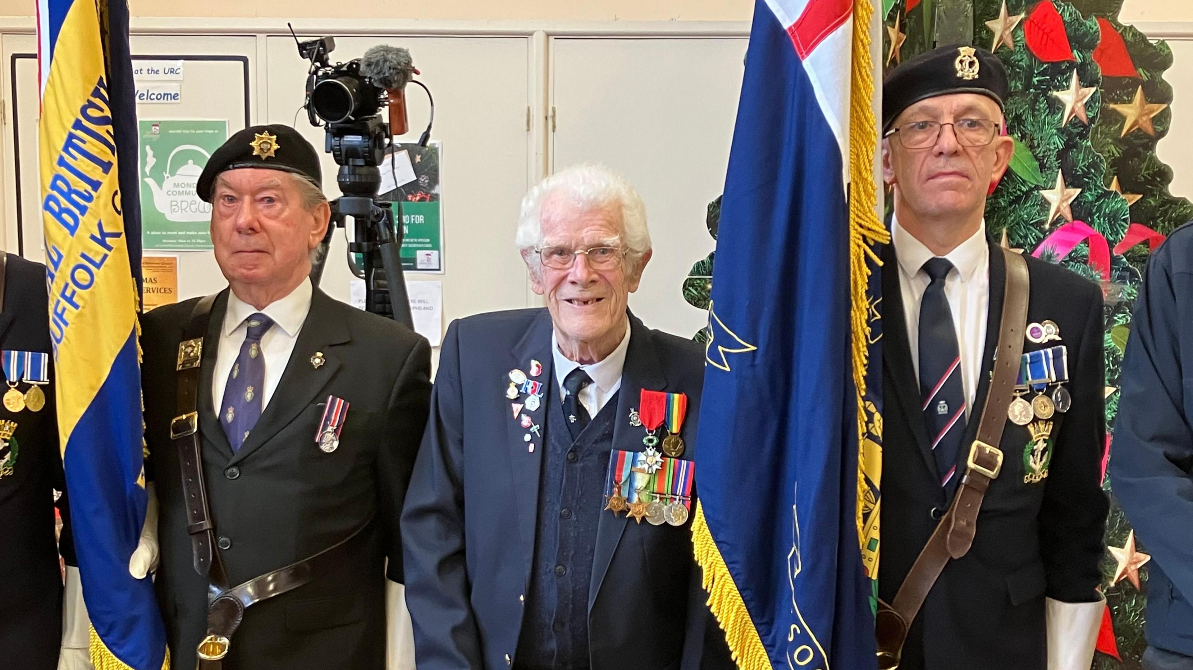 Mr Grant, displaying his medals on his blue jacket, stands between two standard bearers of the Royal British Legion holding flags by their side