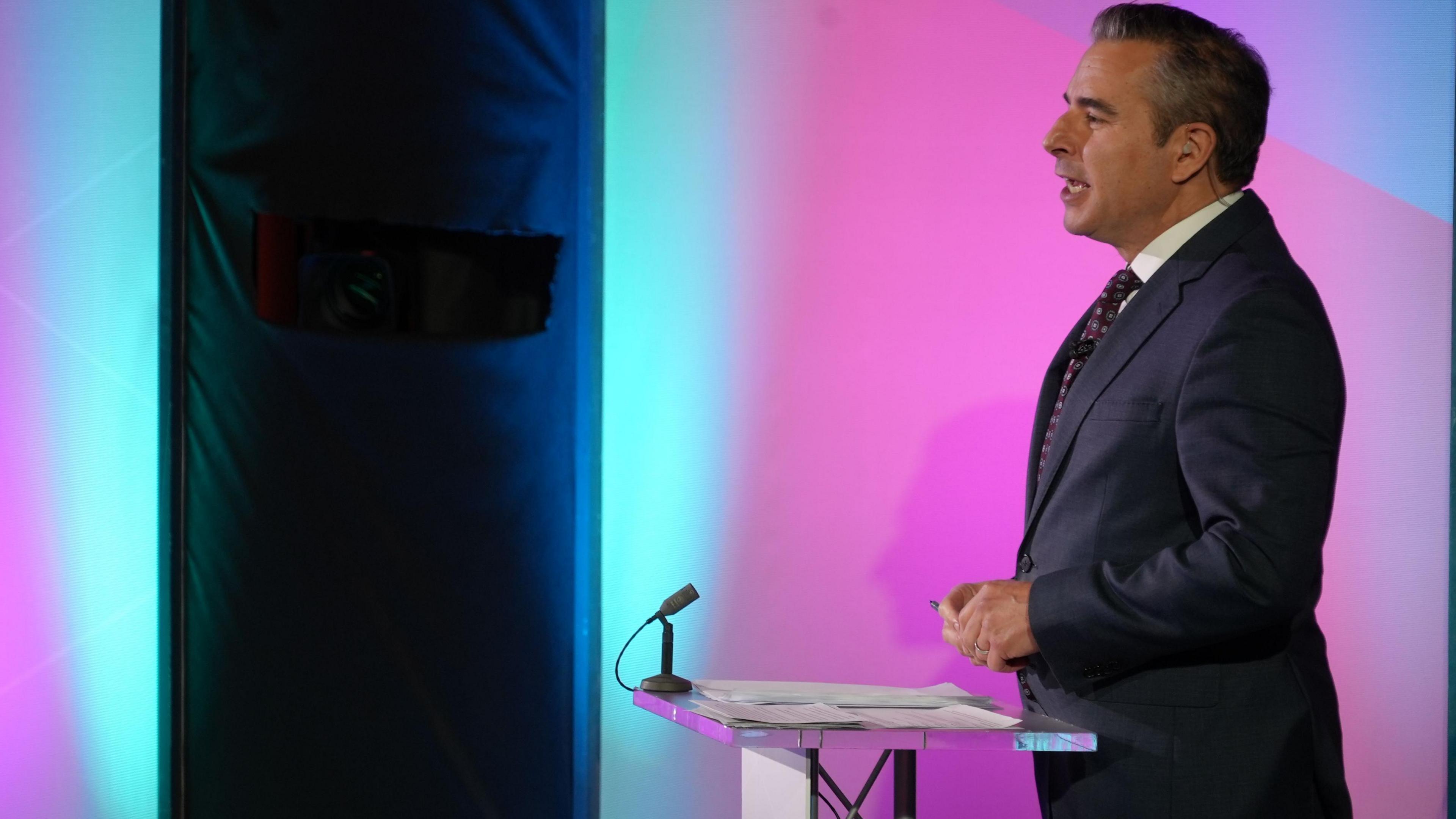 Nick Servini standing at a lectern and speaking, wearing a dark suit and tie. Behind him is a purple and blue set. The lectern has two small piles of notes on it and a small microphone on one of two corners of it furthest away from Nick.