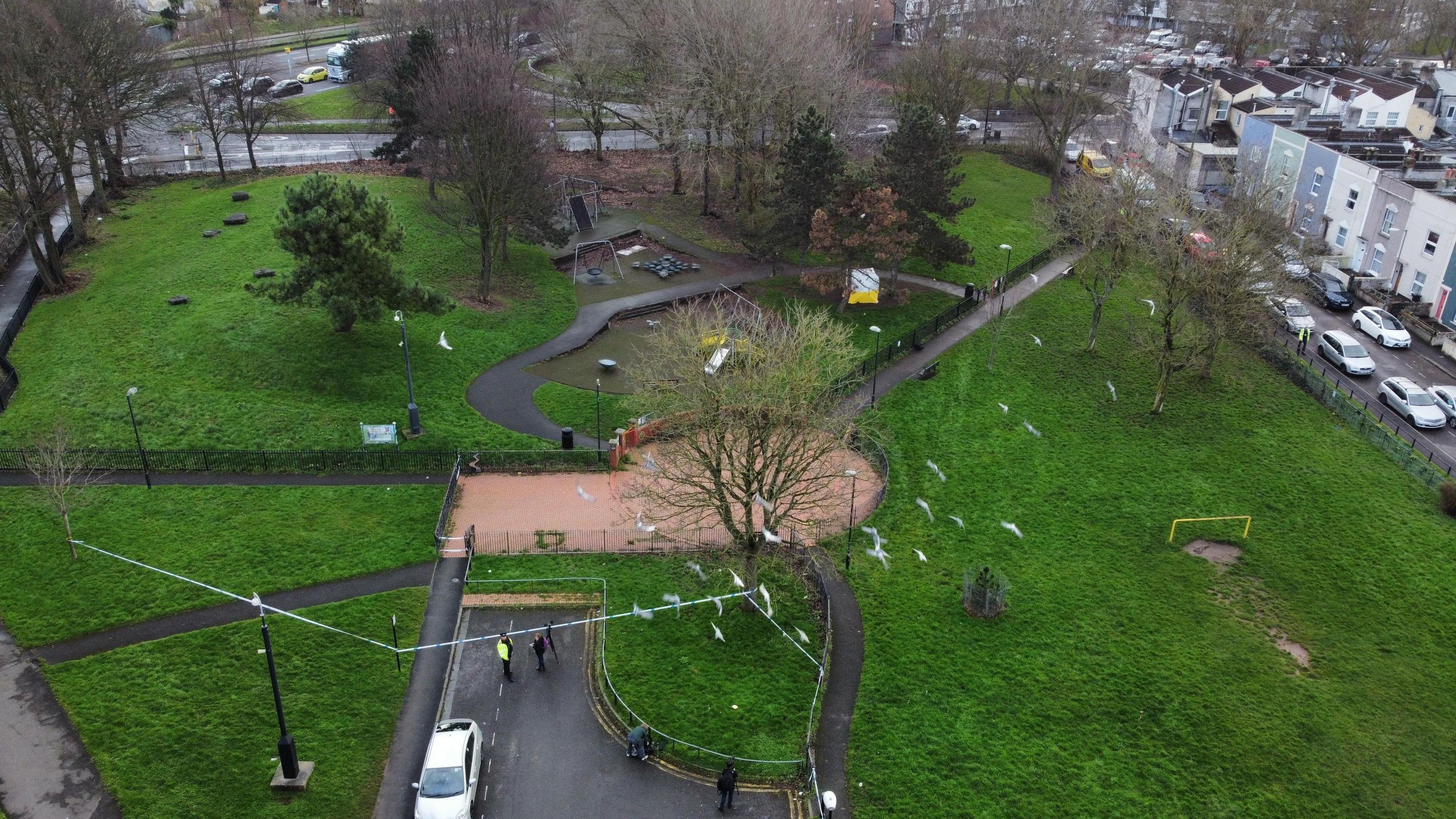 An aerial image of Rawnsley Park. It is a large open green space with paved paths winding through the grass. There is a small play park section where a white and yellow forensic tent has been set up. There is police caution tape securing the area. 