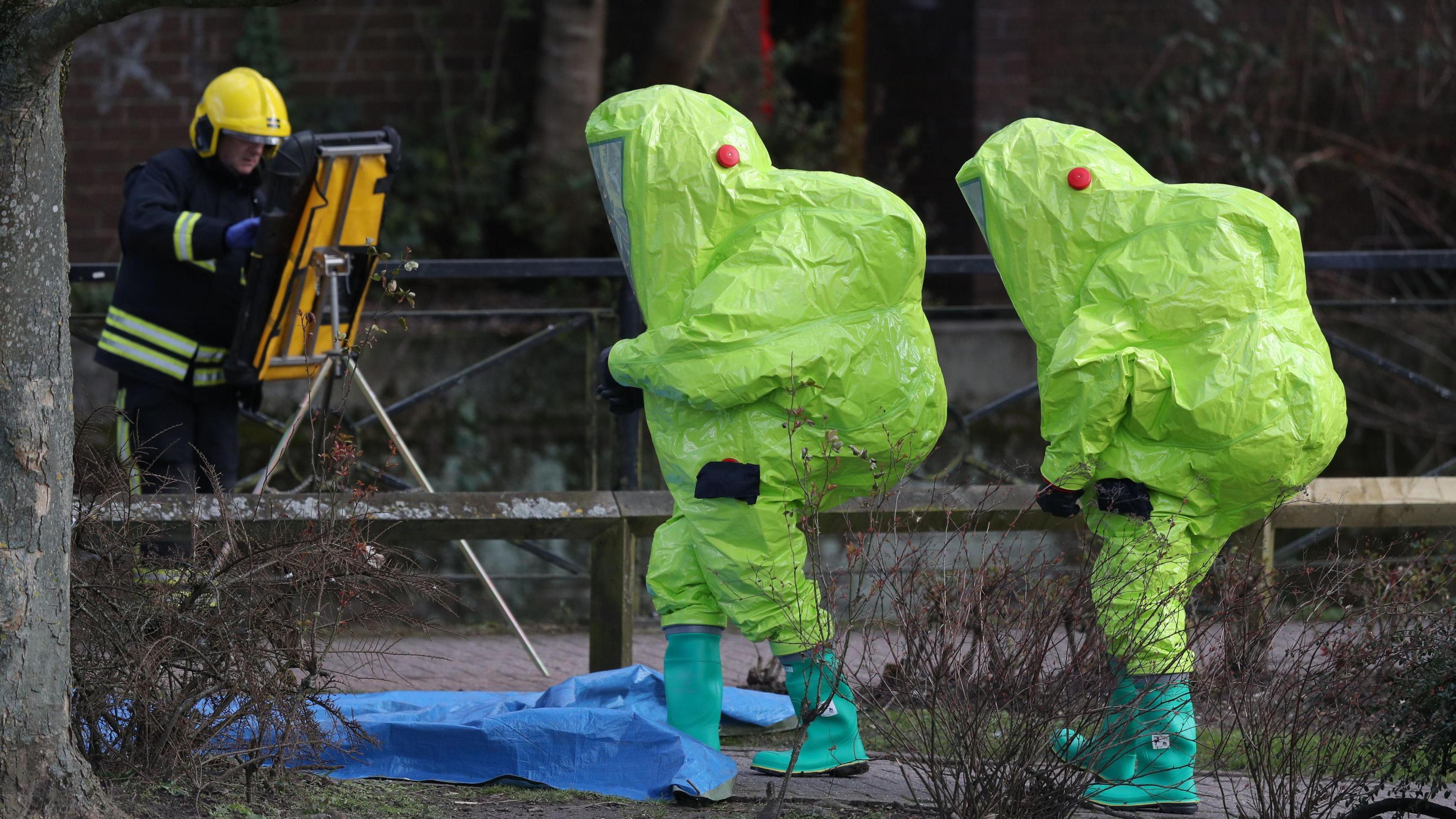 Two people in large neon hazmat suits, yellow. Walking on some pavement in front of a tree on grey day. A firefighter in the background with some equipment.