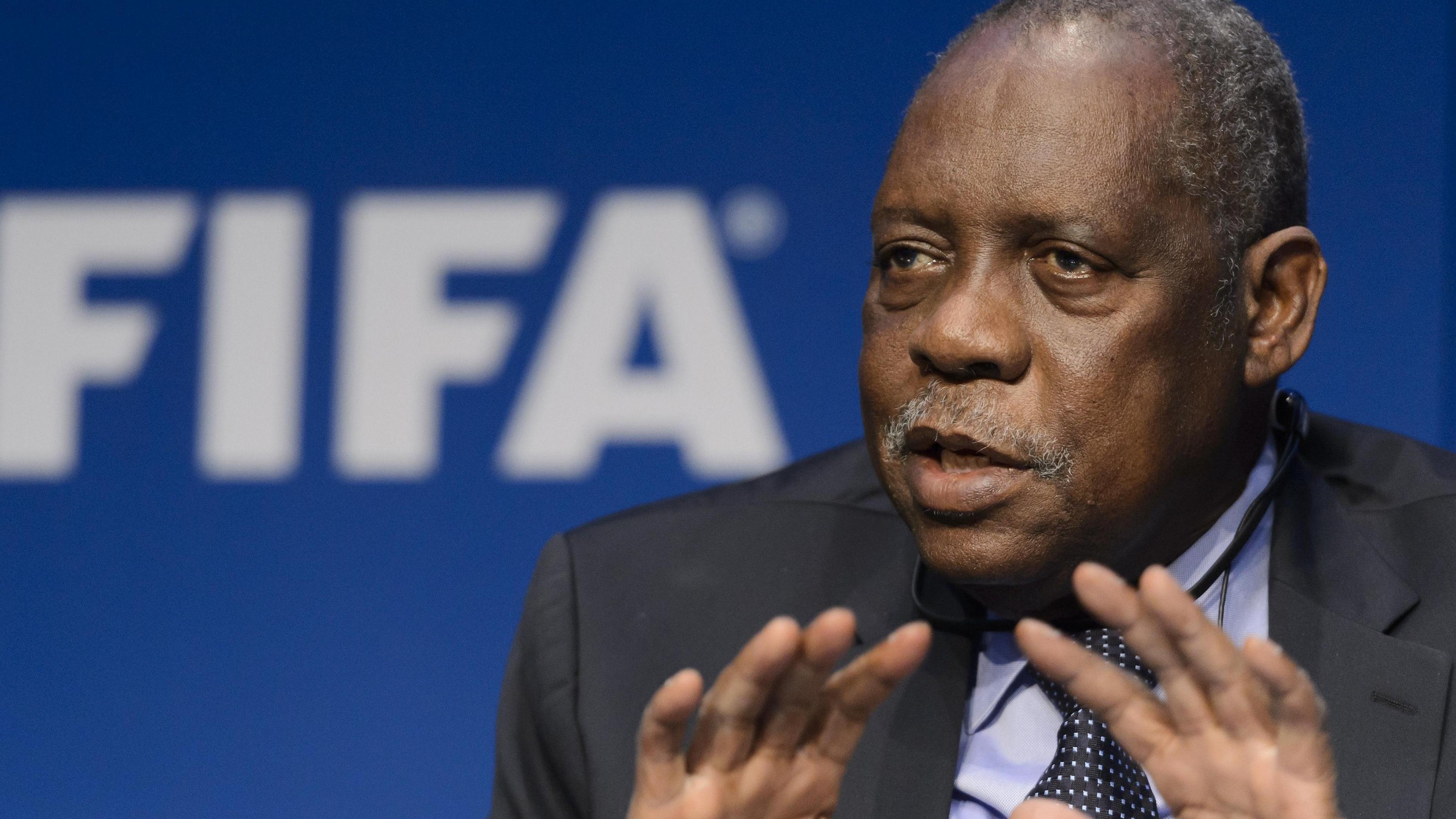 Issa Hayatou, wearing a dark suit, gestures with his hands while speaking in front of a blue wall with a Fifa sign in the background