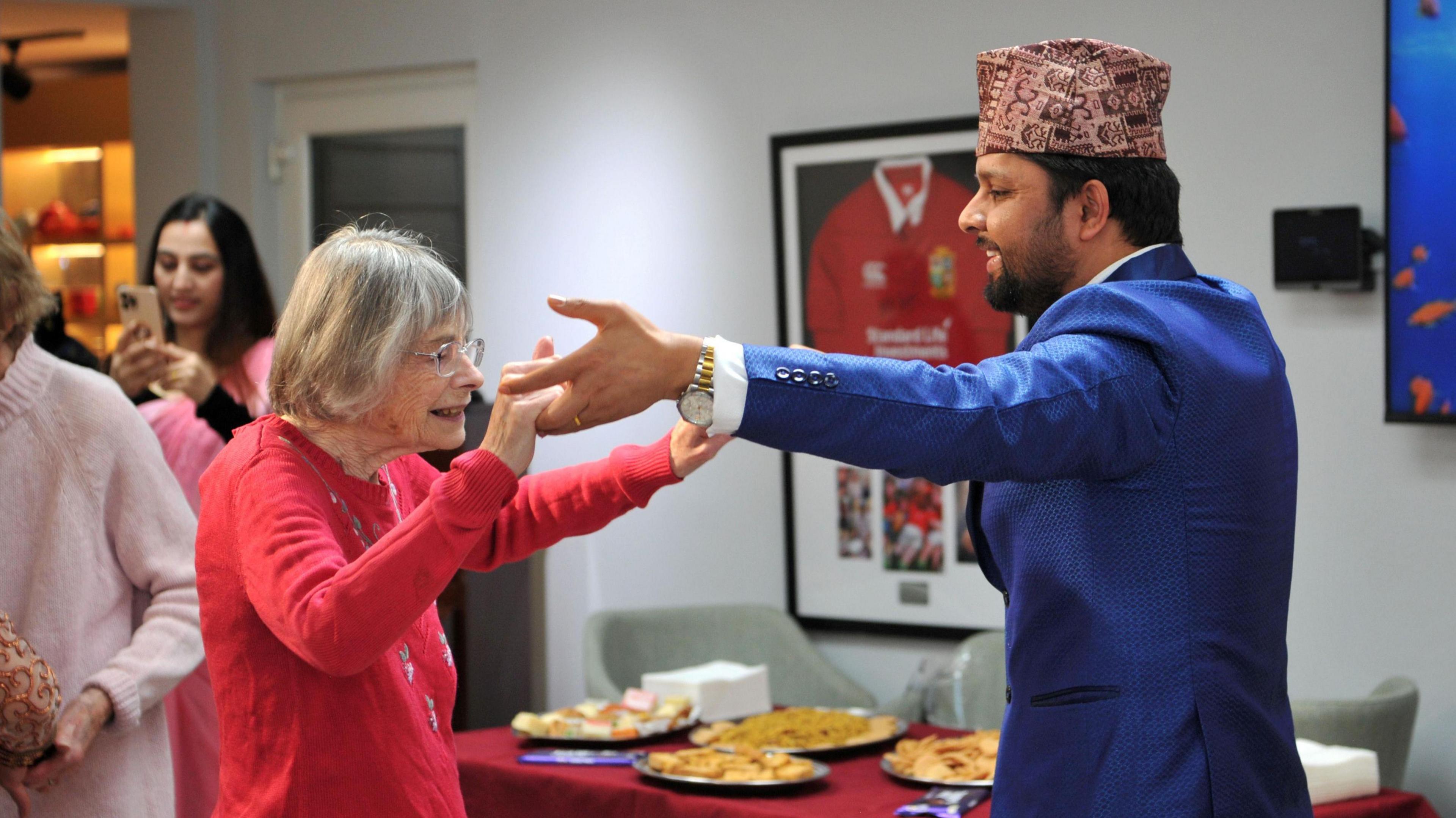 A man is wearing traditional Hindu clothing dancing with a woman who is wearing a red jumper. In the background is traditional food for the Diwali celebration.