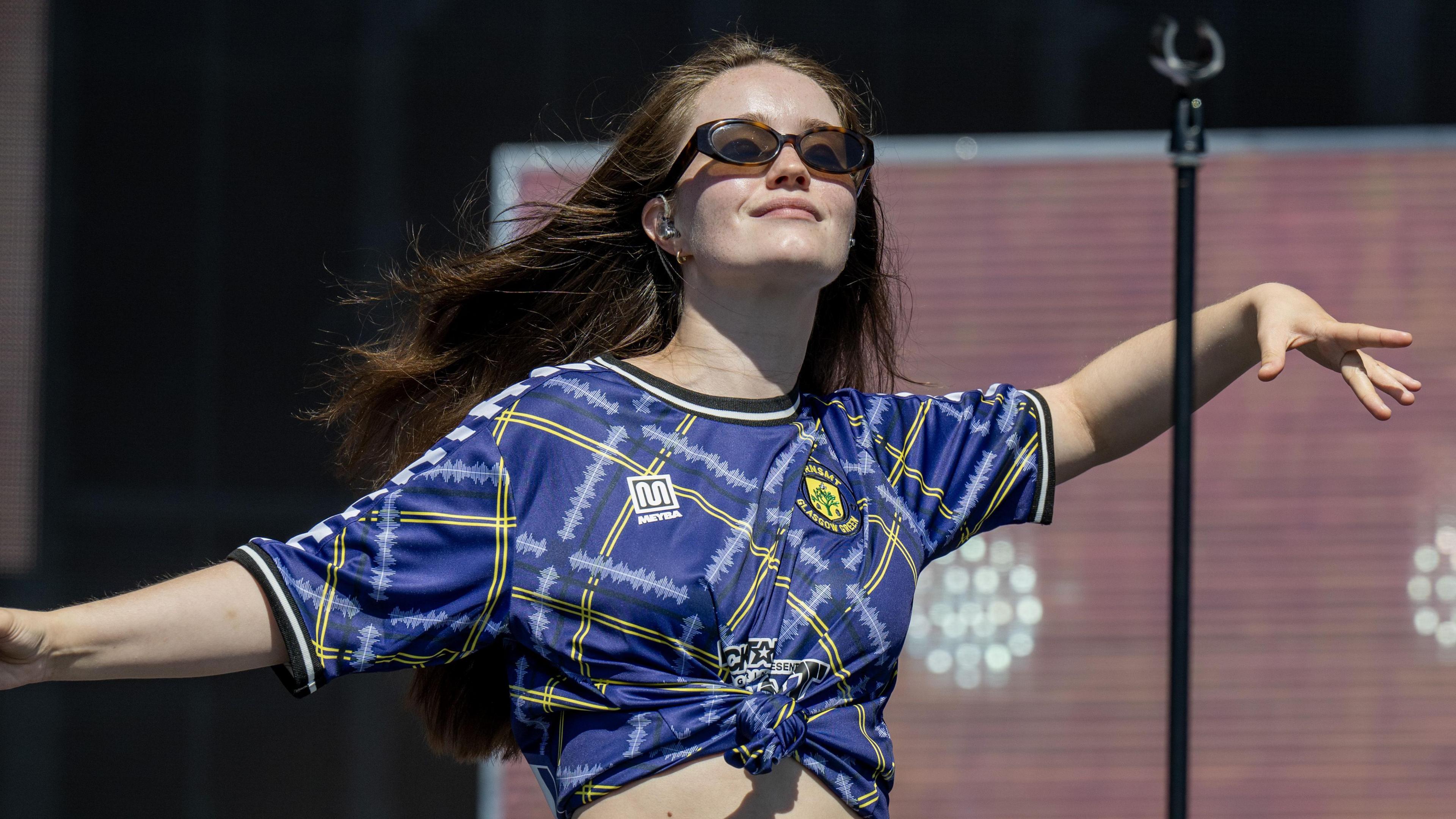 A woman sings onstage while wearing a football top. She has red hair and sunglasses on