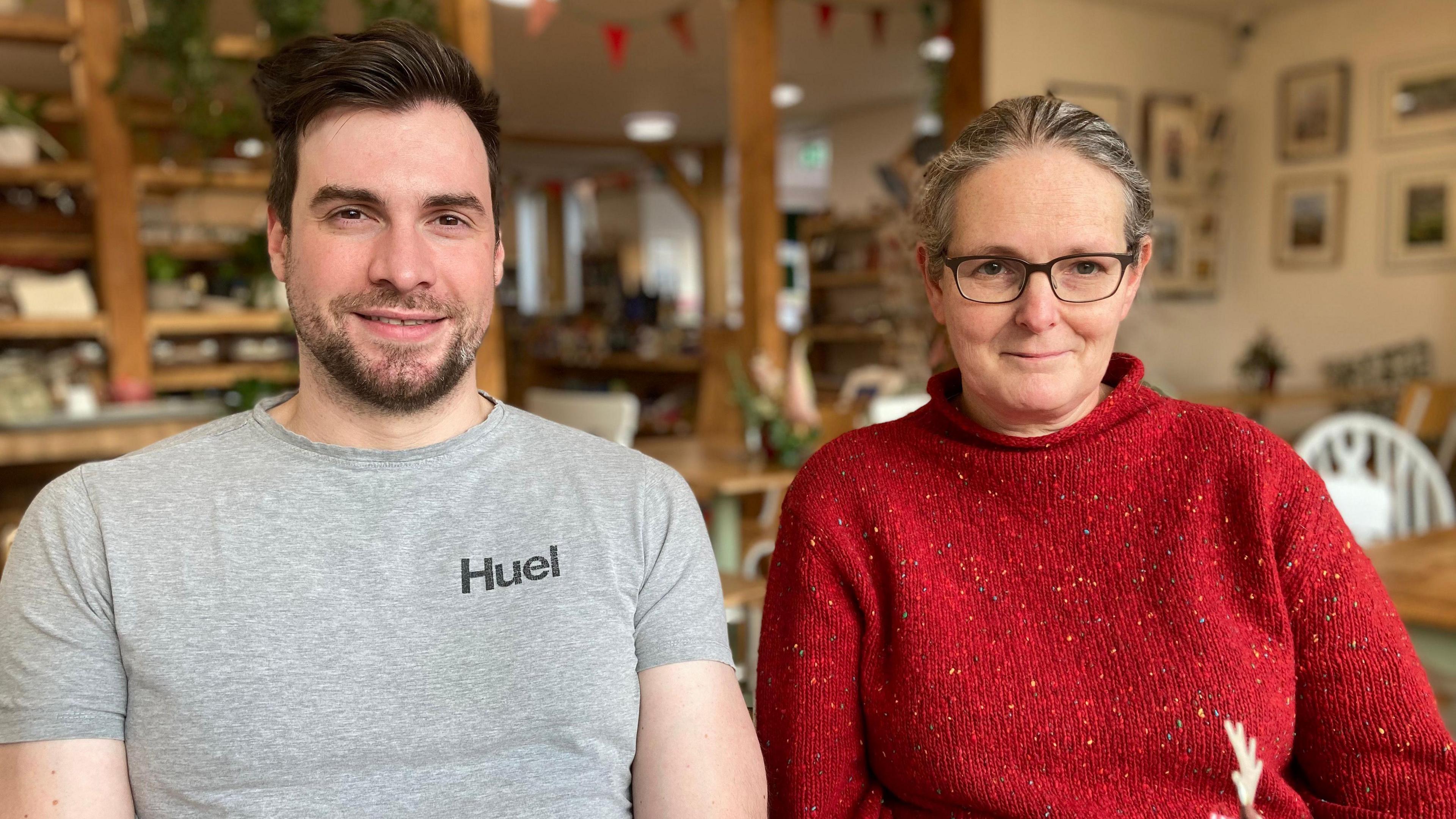 Mr Vaughan-Adkins sitting down at a cafe table on the left. He is wearing a short-sleeved grey t-shirt, has  short dark hair and a beard. Ms Vivian is sitting beside him on the right, wearing black framed glasses and a red turtleneck jumper with colourful specks on it. Behind them you can see the cafe and adjoining shop in the distance.