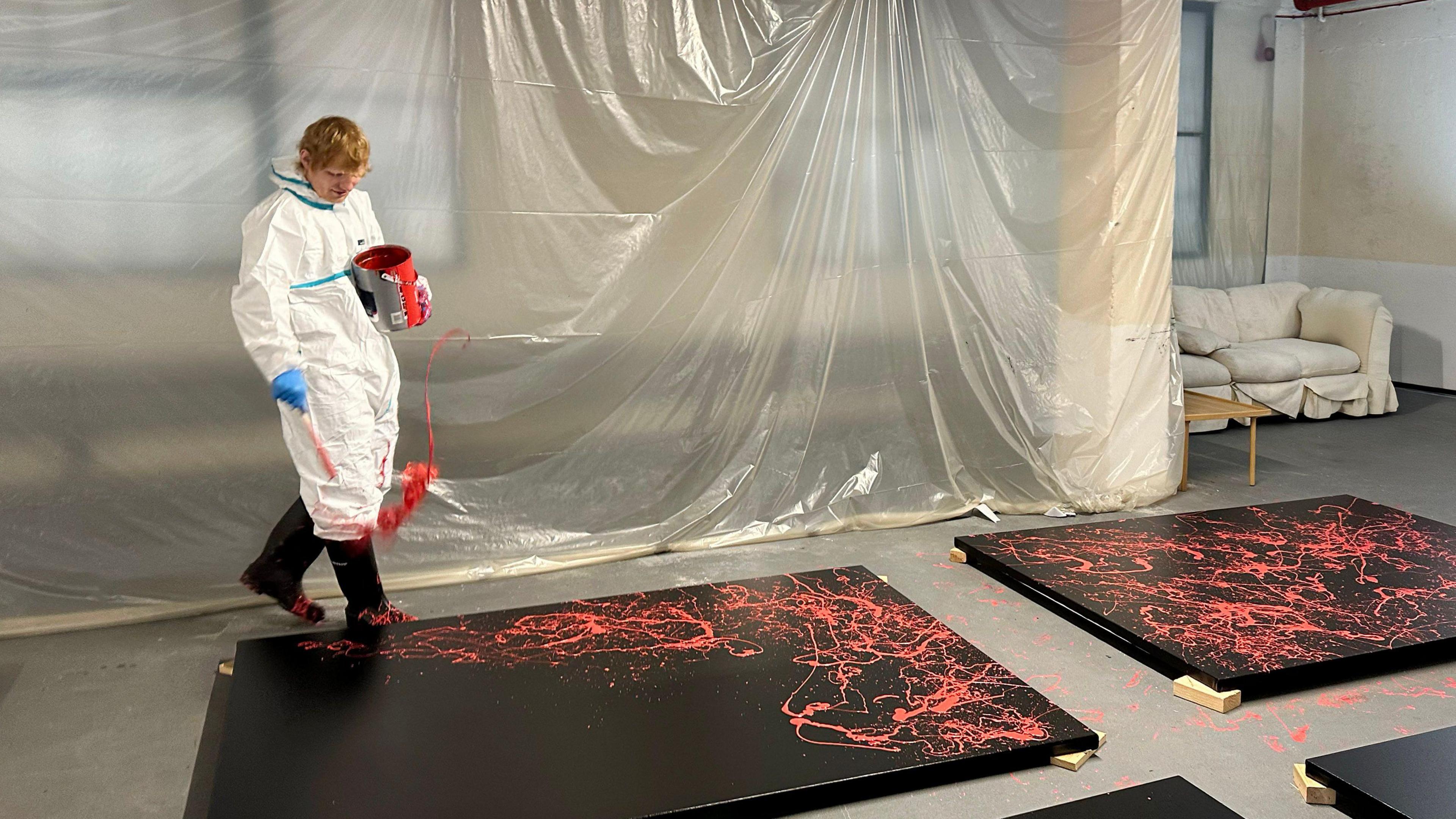 Ed Sheeran walks beside a black canvas that is laid on the floor. He throws pink paint over the canvas. He wears white overalls that are splattered in paint.