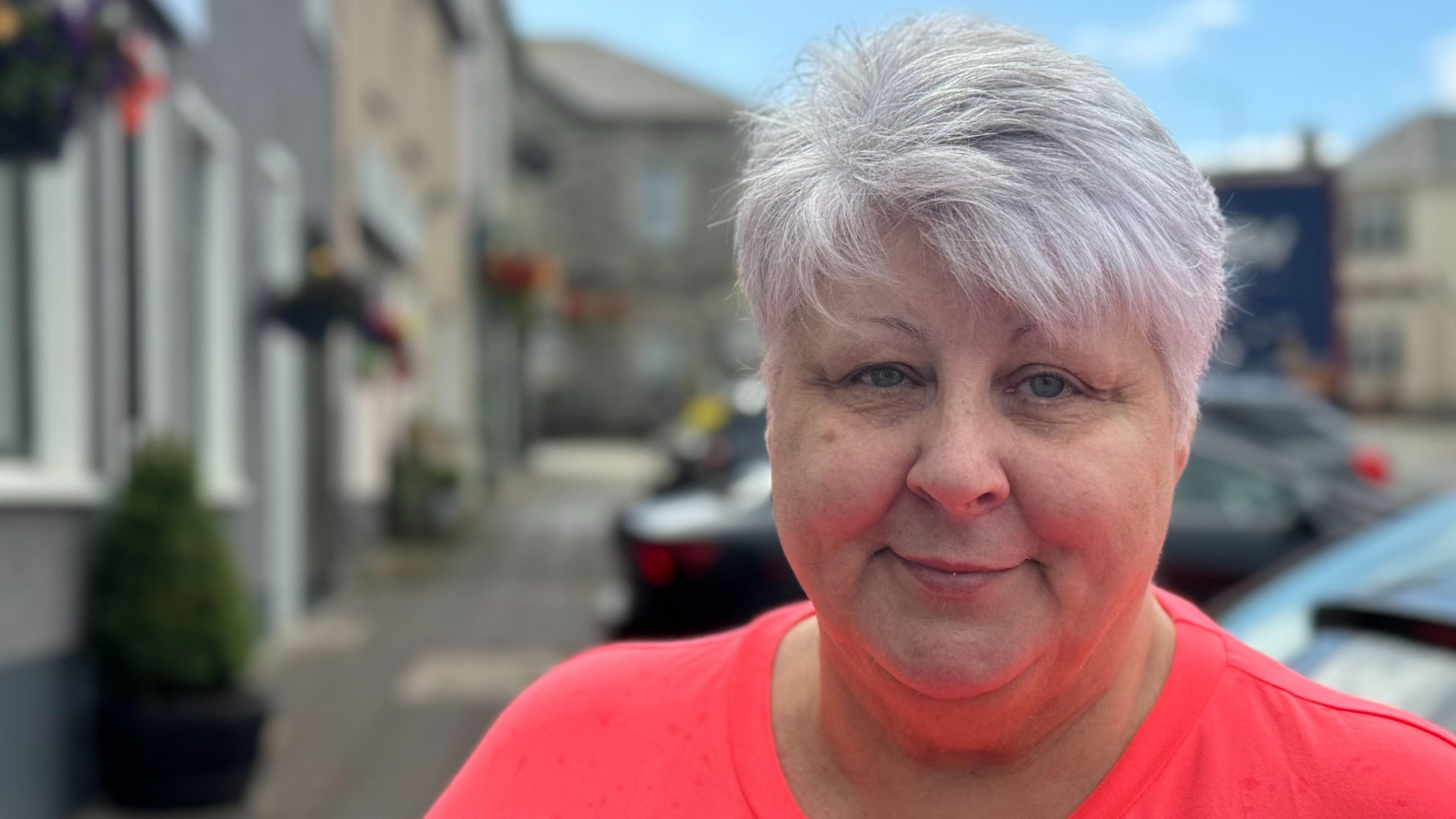 Donna Nugent is standing in the street, looking at the camera. She has grey hair and is wearing a bright pink top.