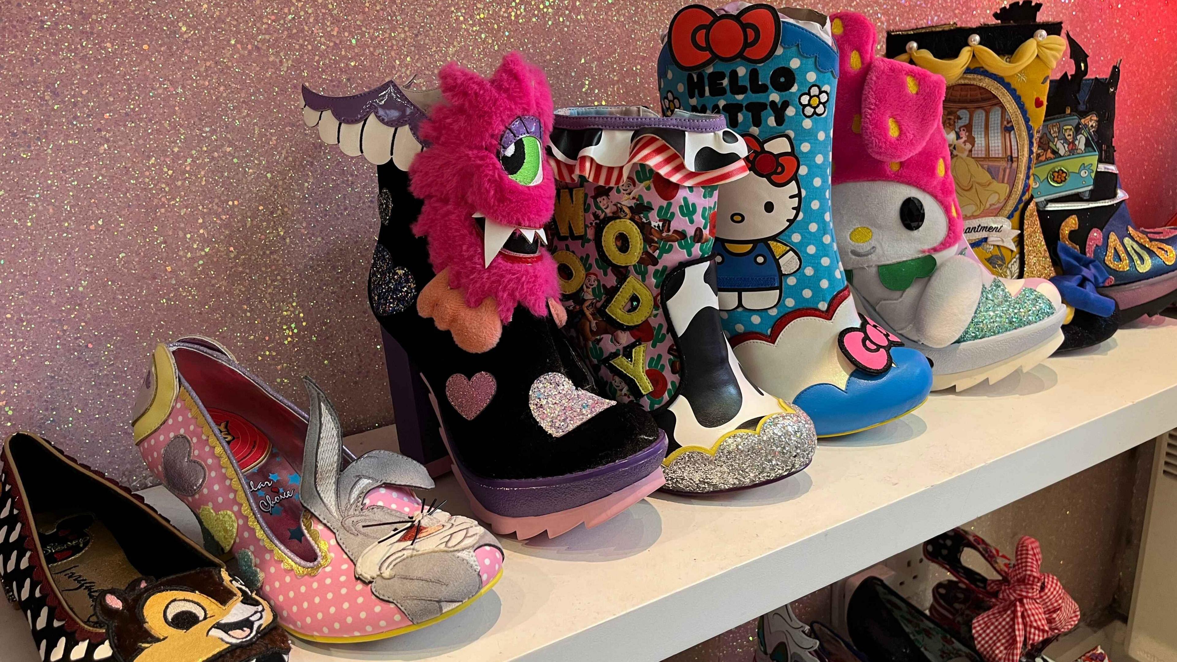 A row of shoes on a shelf which have different characters like Bugs Bunny, Hello Kitty, Scooby Do characters. The wall behind is glittery