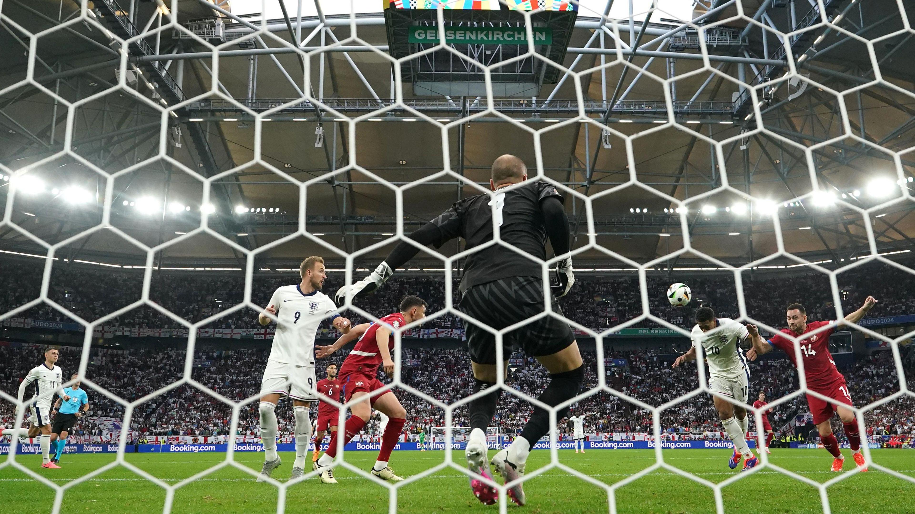 Footballers on a pitch. One heads a ball into a goal past the goalkeeper