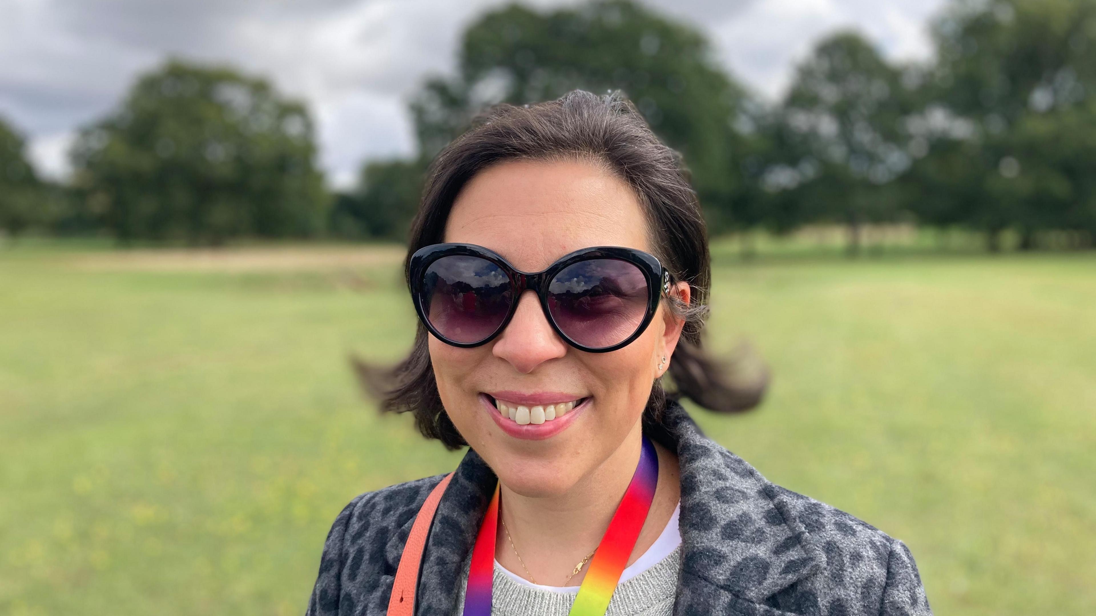 Koulla Jolley smiles directly at the camera as she is photographed outside in a field. She has dark glasses on and is wearing a patterned coat. She is wearing a multi-coloured lanyard and short, dark hair. 