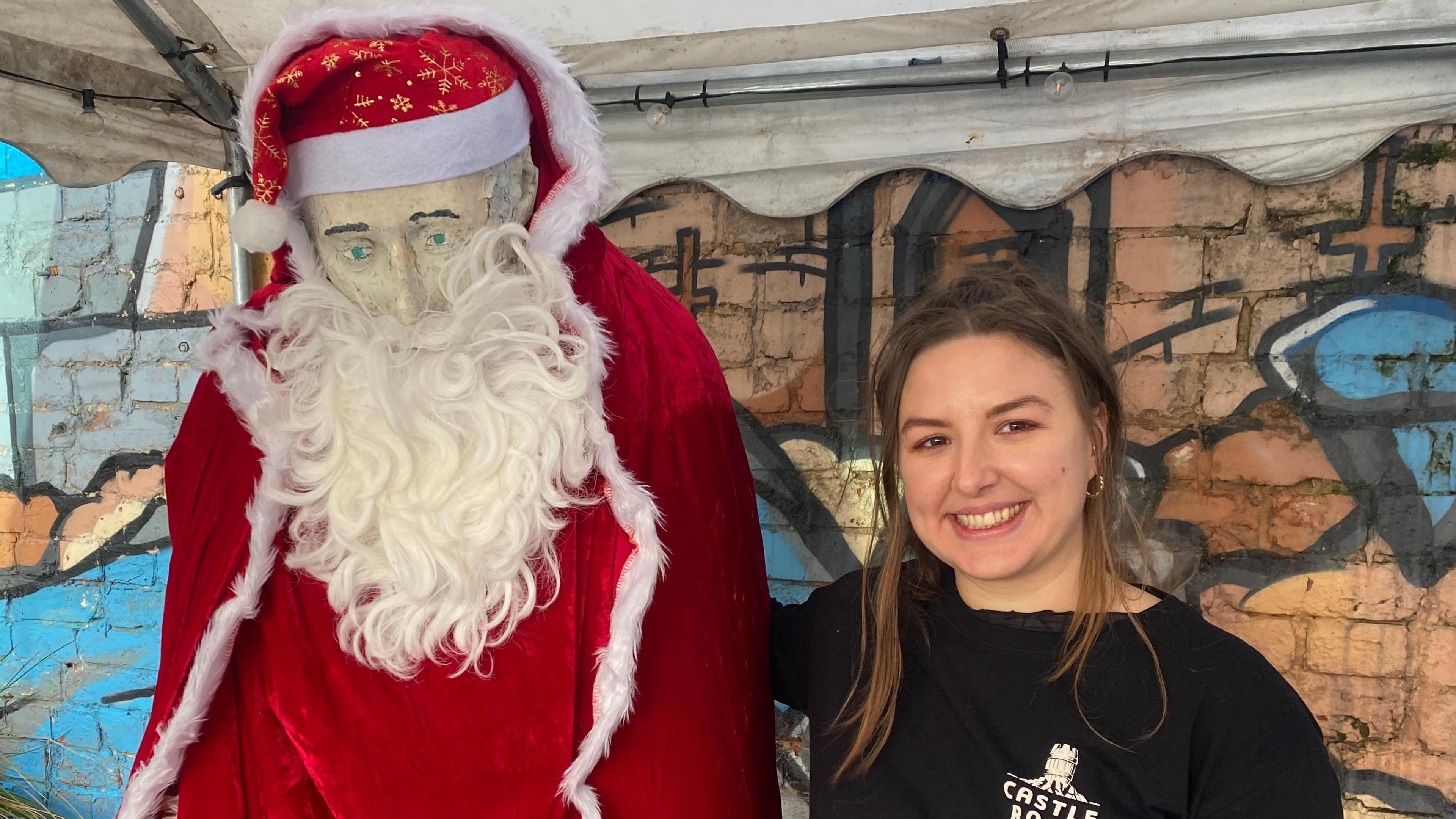 The old general statue wearing a red velvet robe with white fur trim. It has a red Santa hat with gold snowflakes on its head. It is also wearing a long white beard. A woman, dressed in a black top has her arm around it. She's smiling.