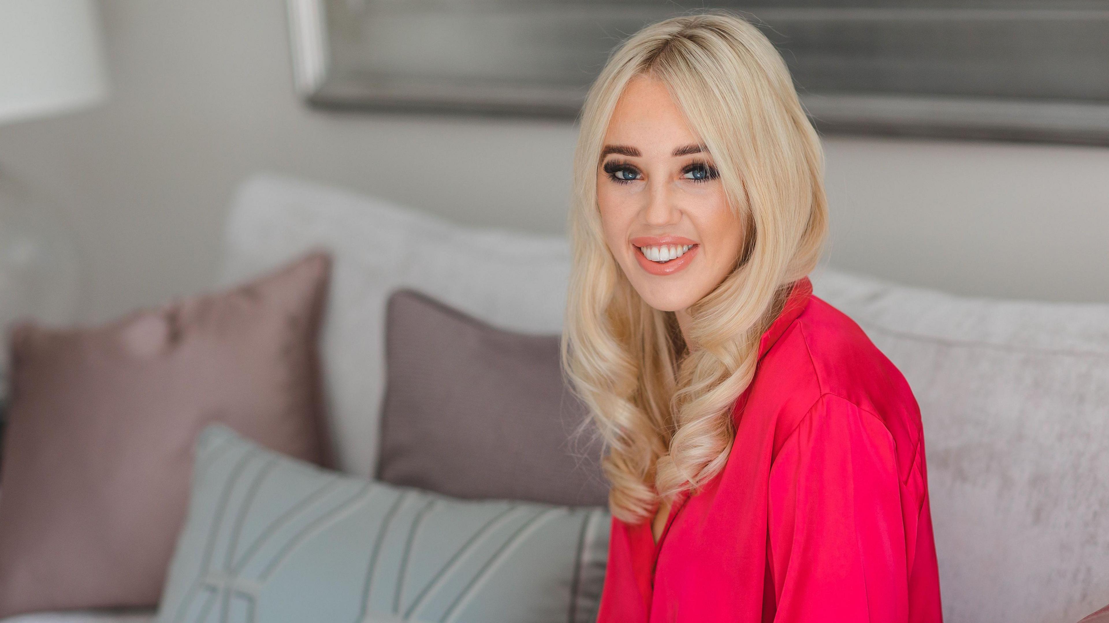 Emma sits on a pink couch and smiles at the camera. She has long blonde hair and wears a pink shirt.