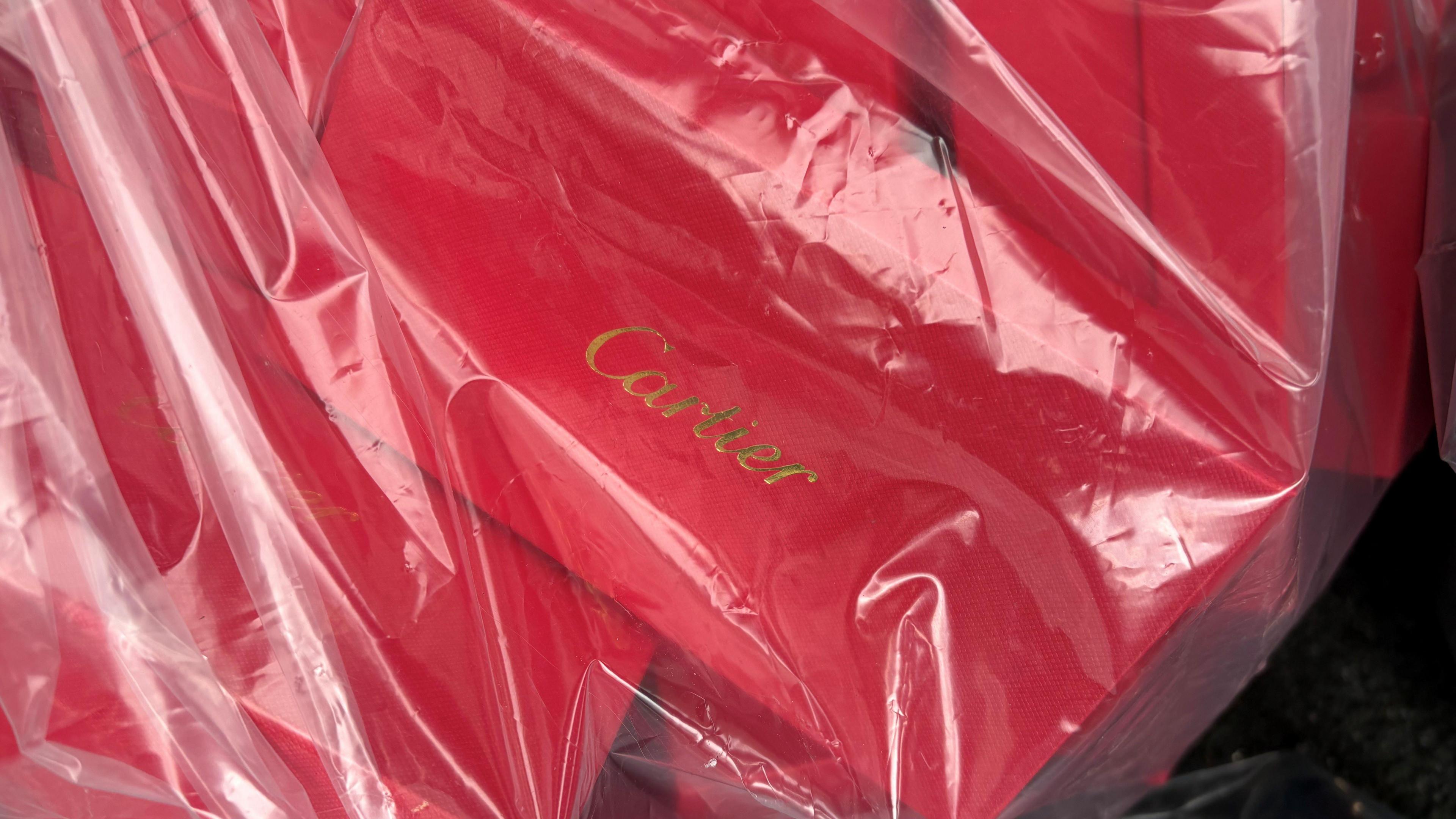 A close-up image of a red box with the brand Cartier written in gold. It is among a number of similar looking boxes in a clear plastic bag.