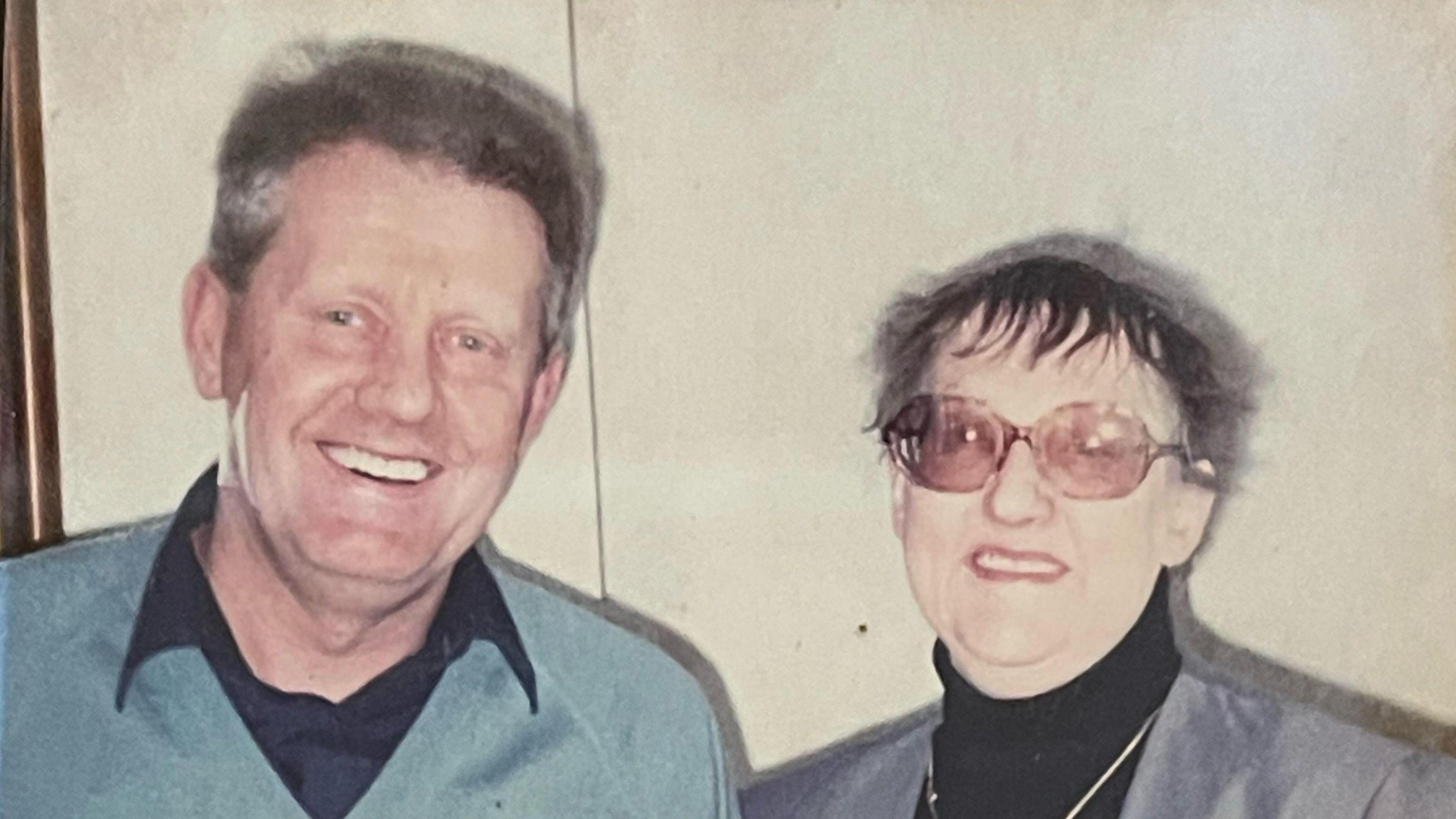 Two people in an old photo - on the left is a man with brown hair in a blue jumper and black shirt. On the right is Patricia, a woman with a black roll neck, a grey blazer and glasses.