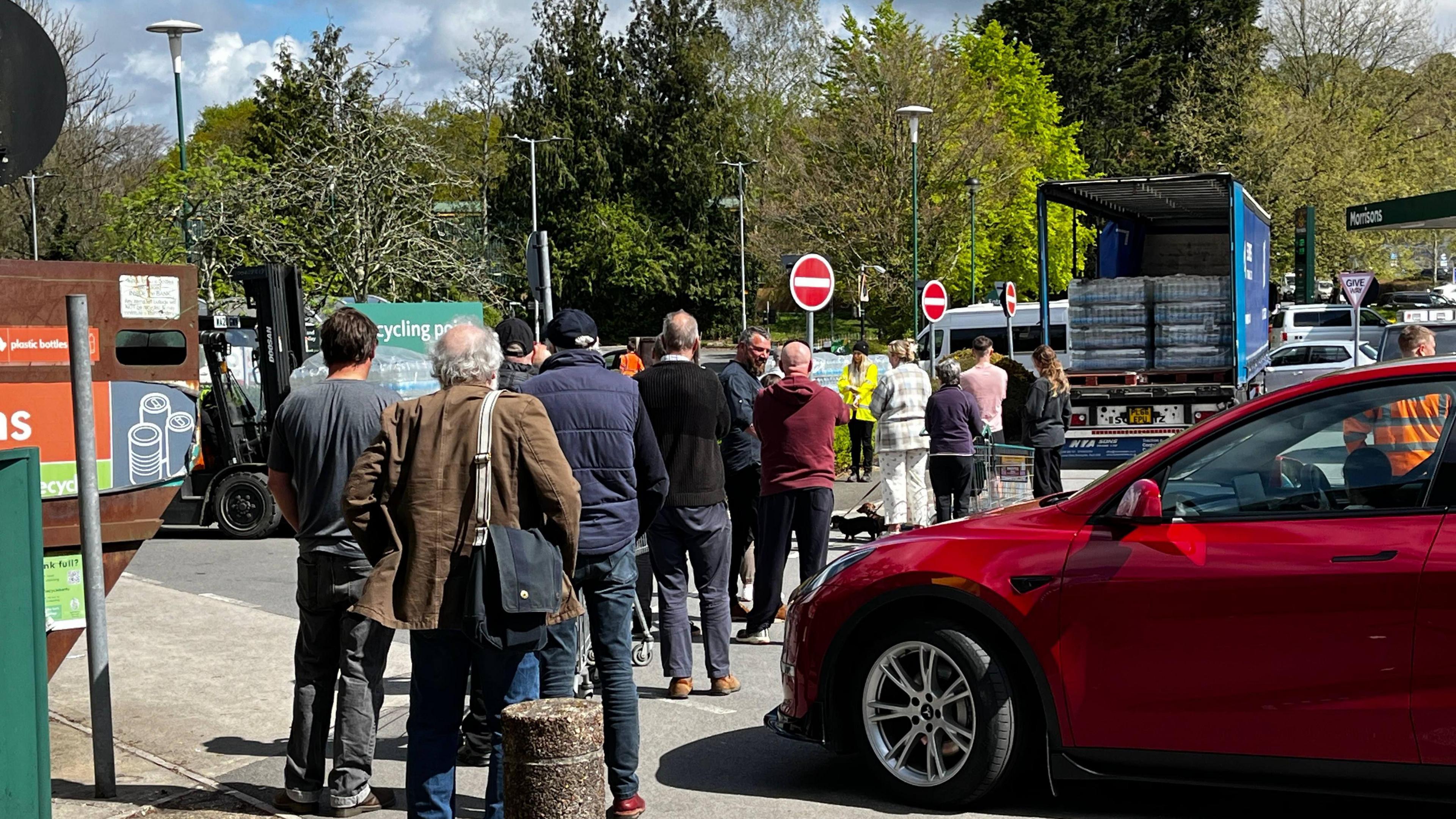 People queuing at a water station in Tavistock. There is a lorry filled with crates of bottled water in the distance with a person wearing hi-vis stood next to it. On the right, there is a parked red car.