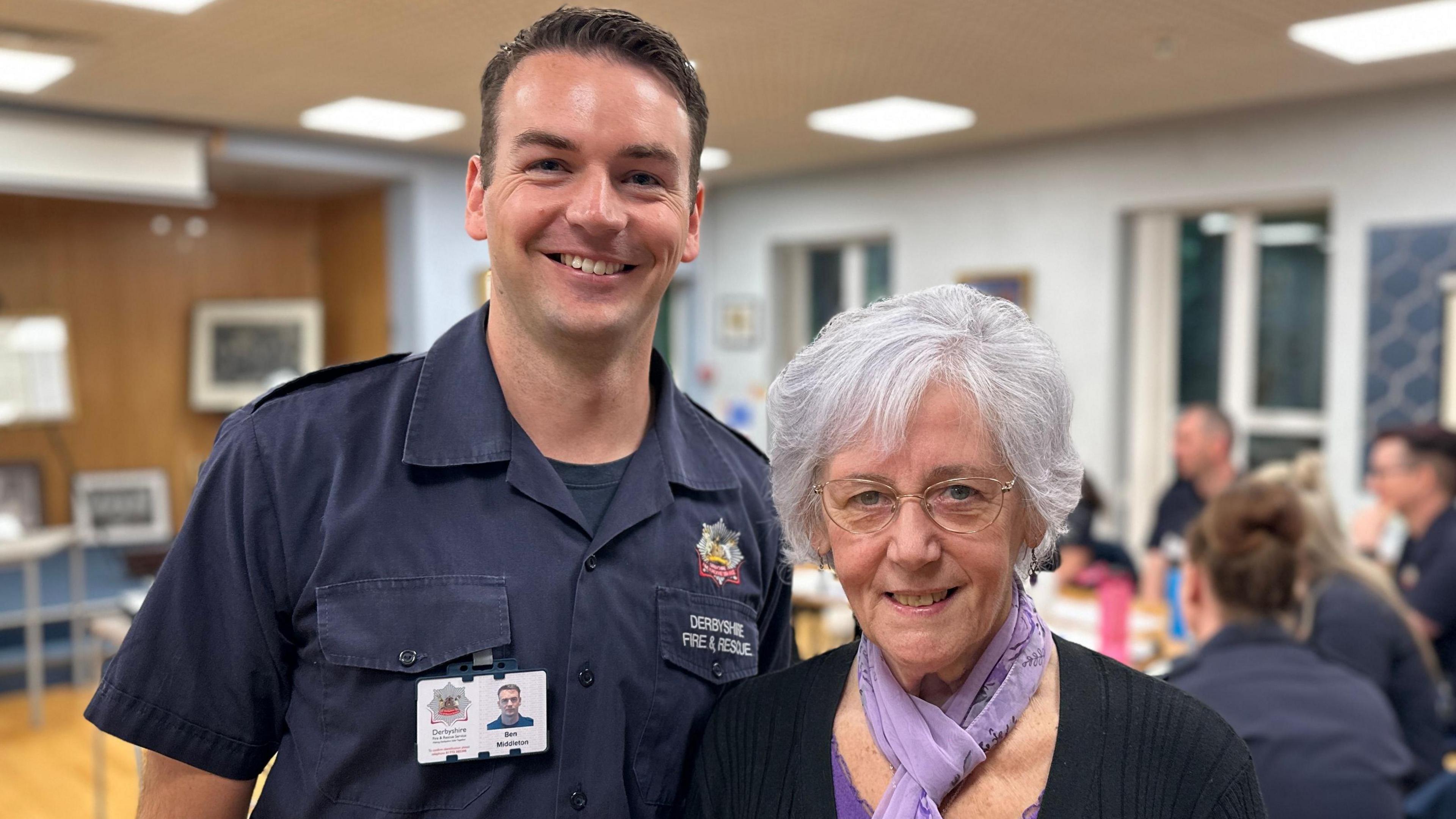 June Lush pictured with firefighter Ben Middleton. Ben has short brown hair and is standing next to June, wearing a blue polo top with a name badge, while June has grey hair, and is wearing a purple scarf. They are stood in front of a group of emergency services staff in sign language training