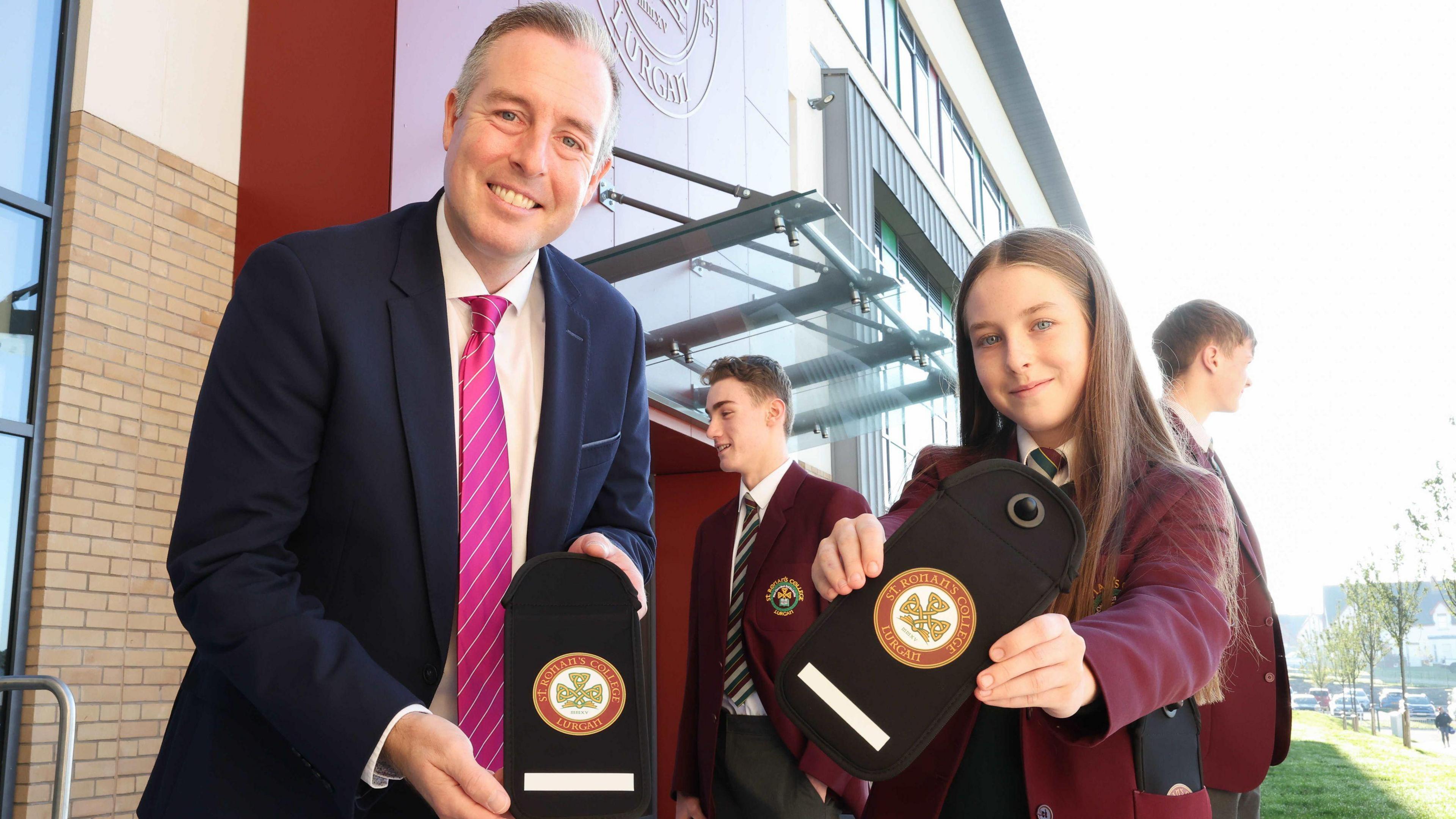 A man in a navy suit, white shirt and pink tie holds a black pouch with a red and white circle badge on it. A young girl stands beside him holding an identical pouch, they both smile to the camera. The girl wears a red blazer with a white shirt and green, red and grey striped tie. Behind the pair are two boys wearing the same blazer shirt and tie combo. Behind all four is a red brick building with rows of clear windows and a large red entrance. 