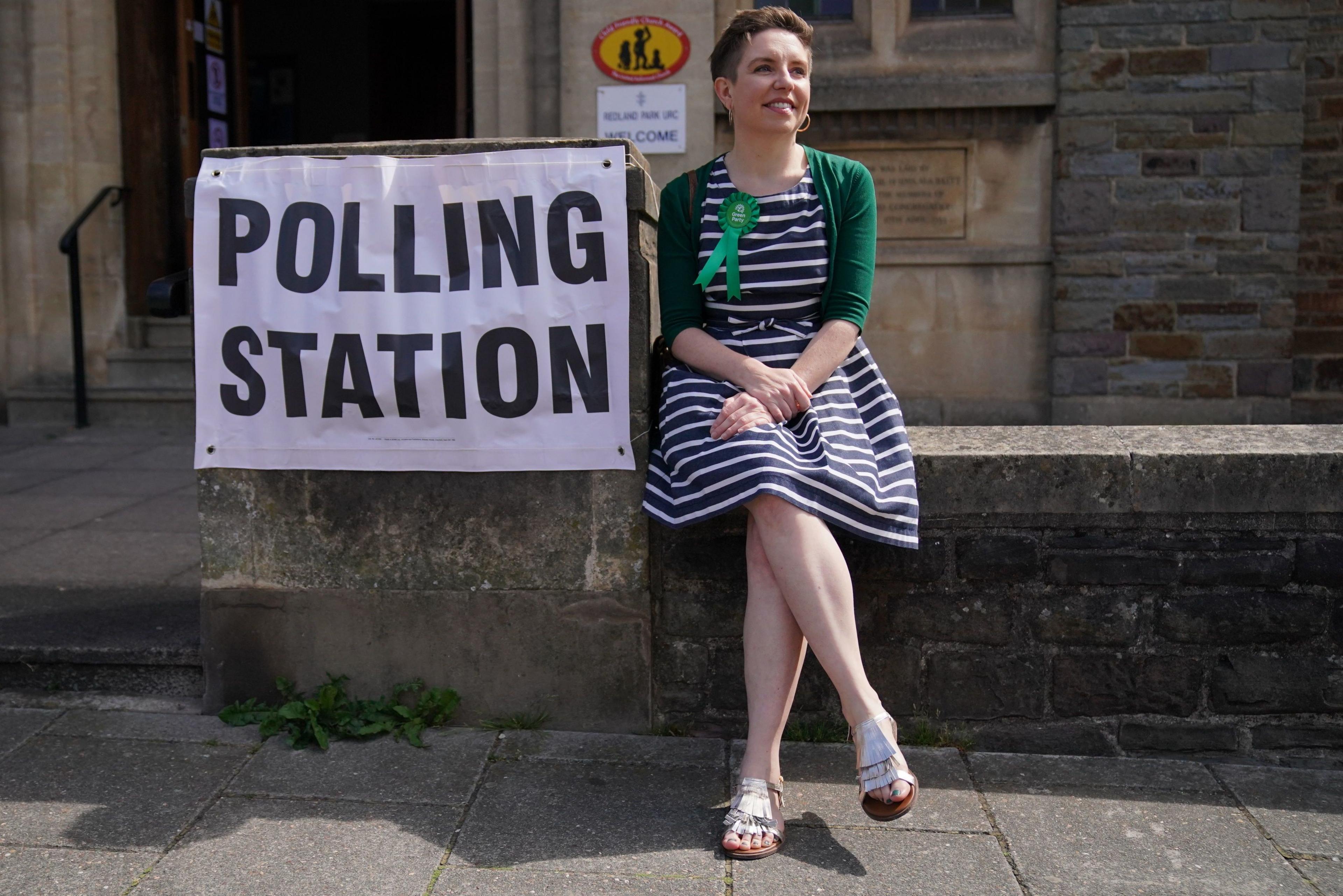 Bristol MP Carla Denyer sits on a wall by a 'Polling Station' banner wearing a green rosette.