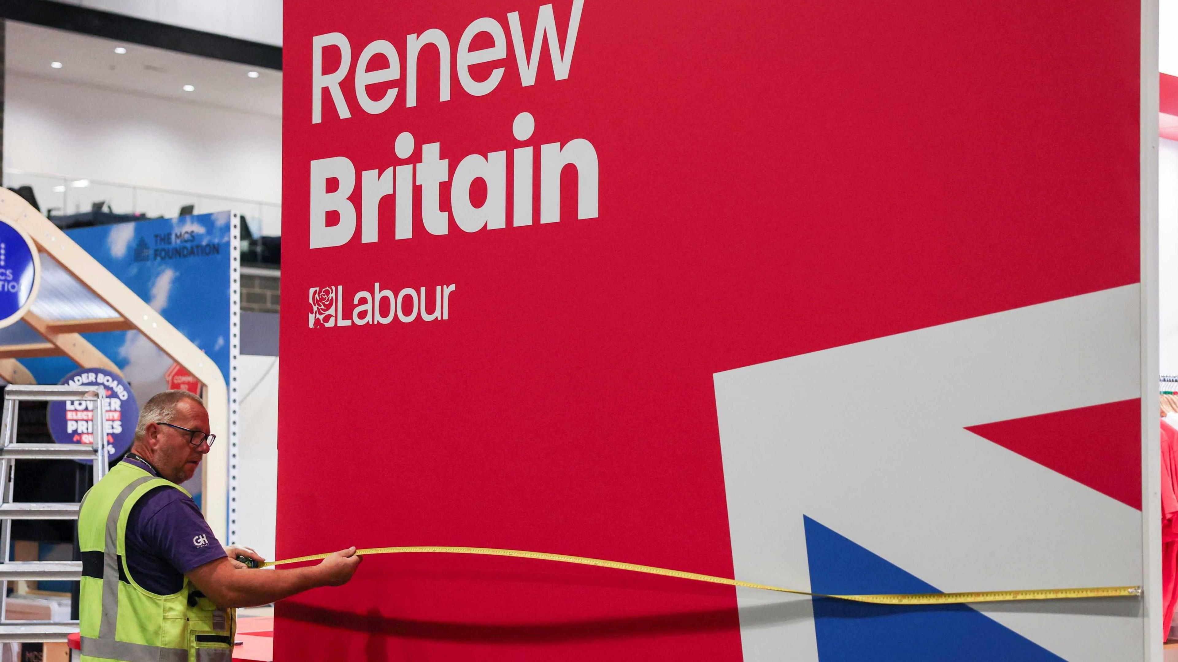 A worker in high vis tabard is standing by a sign saying Labour - Renew Britain over a union jack - he has his measuring tape out and extended the length of the sign. 