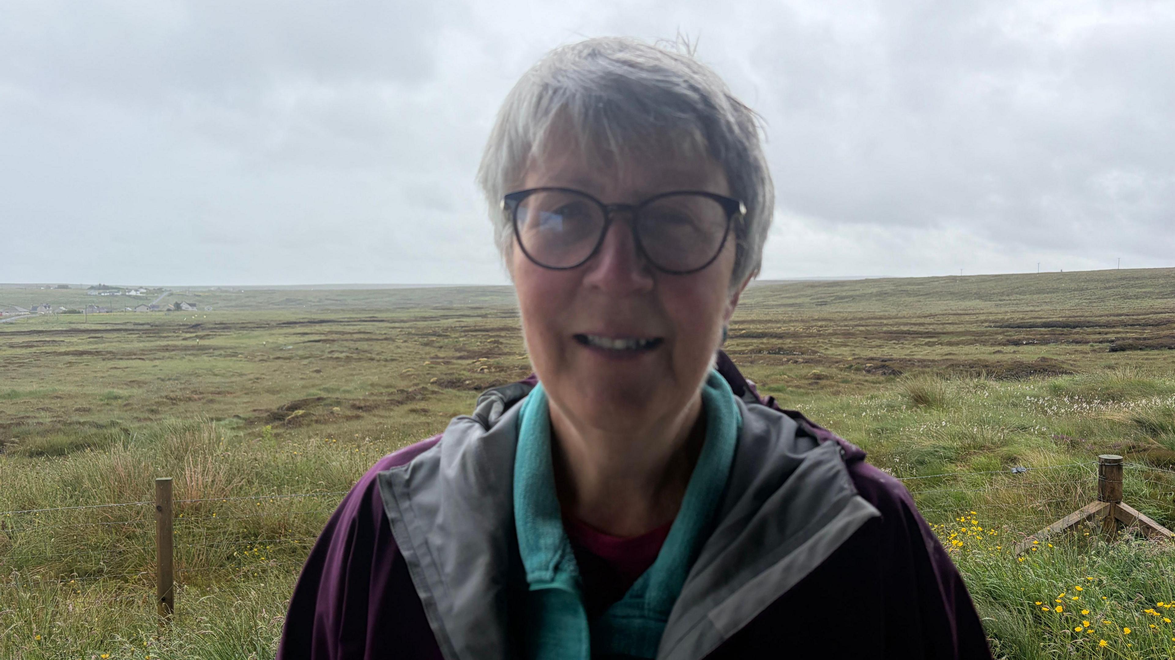 Boireannach le falt goirid, liath agus speuclairean na seasamh air a' mhòinteach.