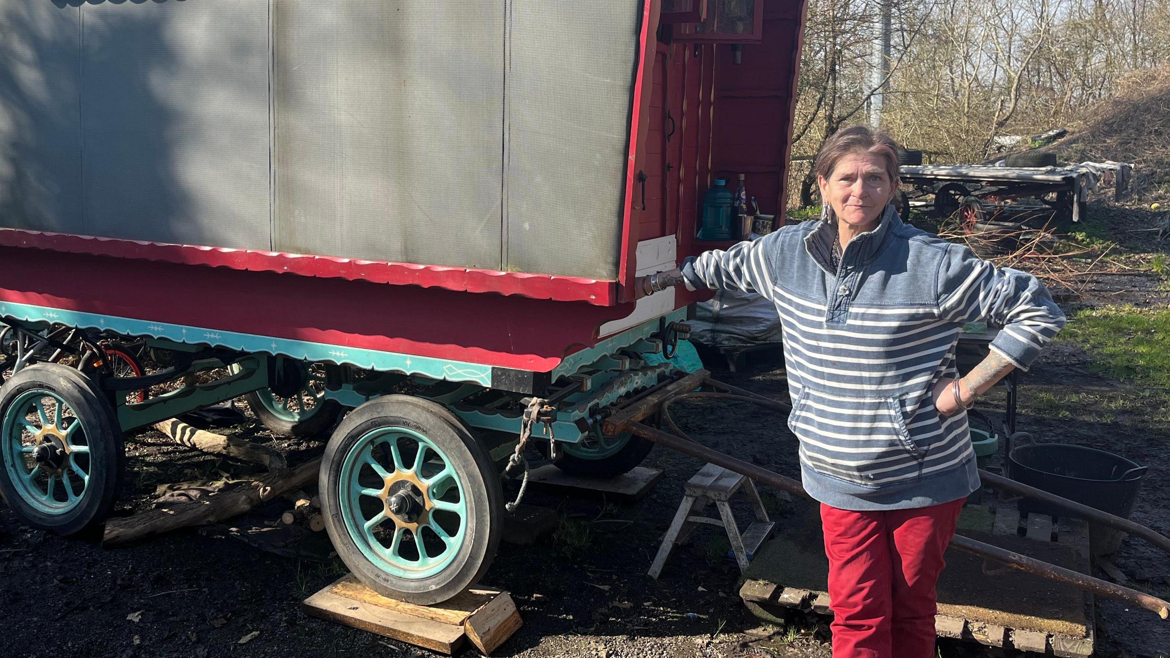 Rose Brash looks at the camera in a stripy jumper and red trousers, leaning against a traditional horse-drawn caravan