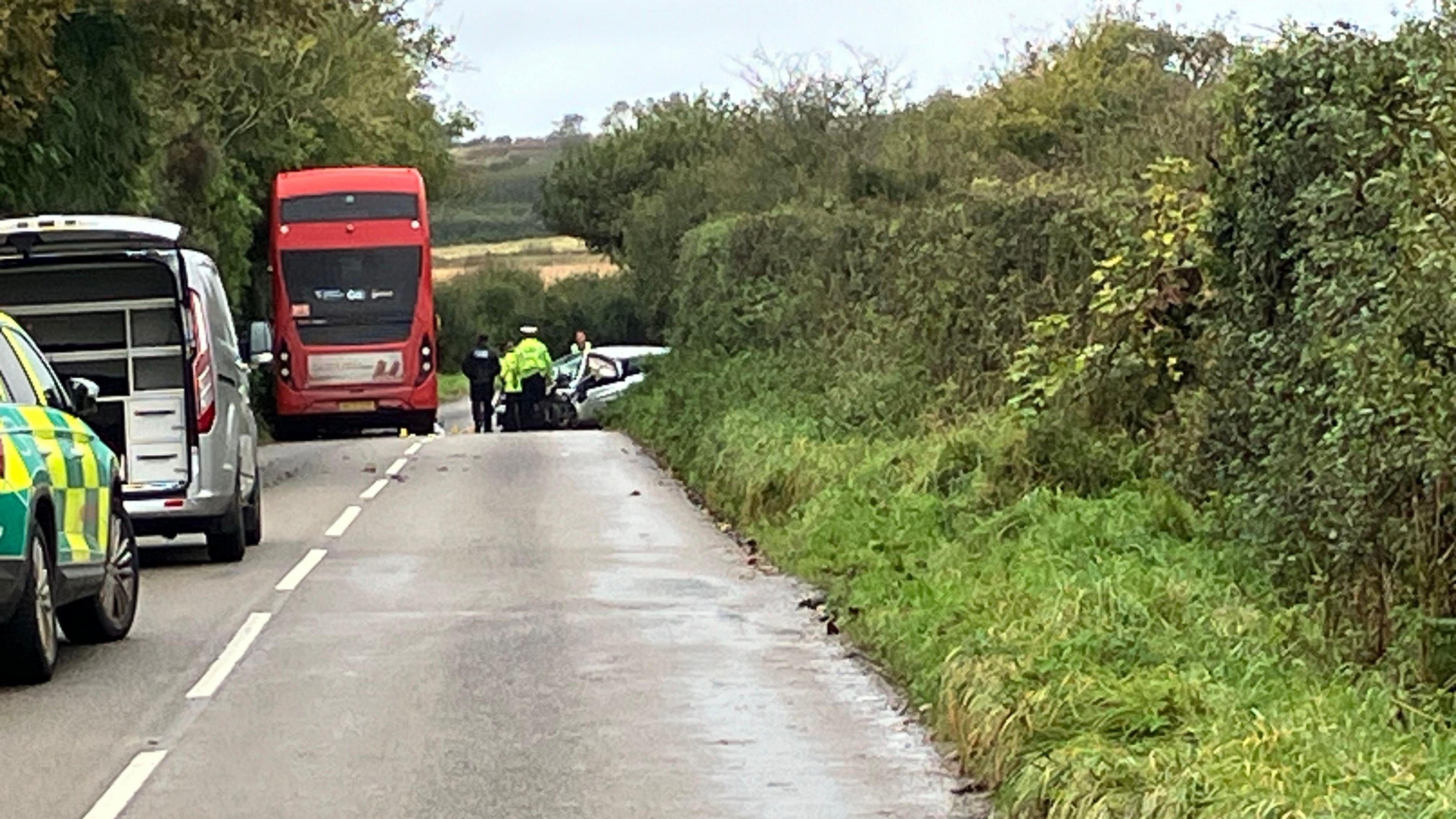 A red double decker bus on the left-hand side of the road with police officers between the bus and car. One officer is in a black jacket and two are wearing yellow hi-vis jackets. On the right, in the green hedges, is a damaged silver vehicle. On either side of the road is grass and hedges. Behind the bus on the left is a green and yellow ambulance vehicle and a silver van.