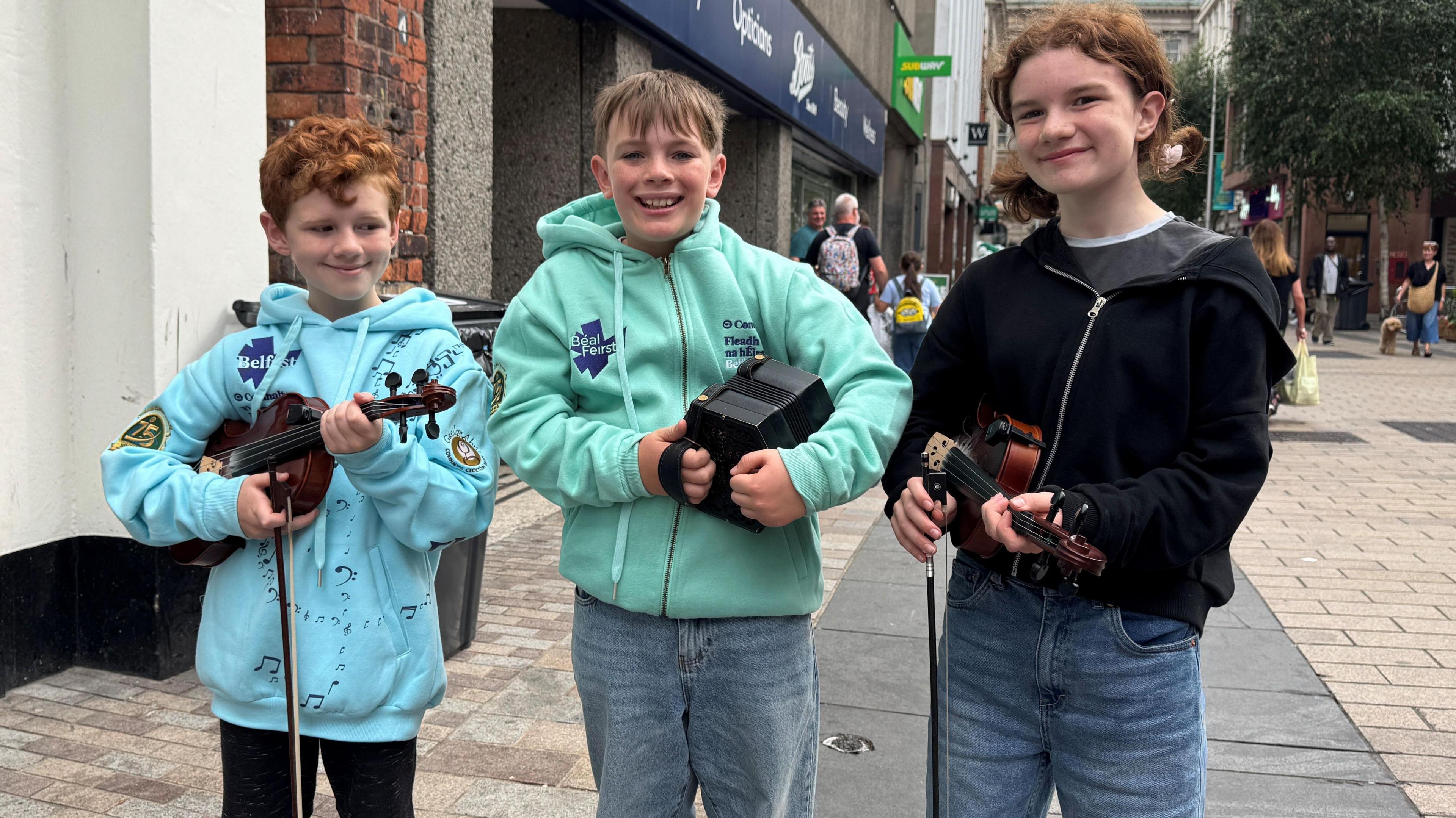 Three children stand together holding instruments, two have red hair the middle one has brown hair.