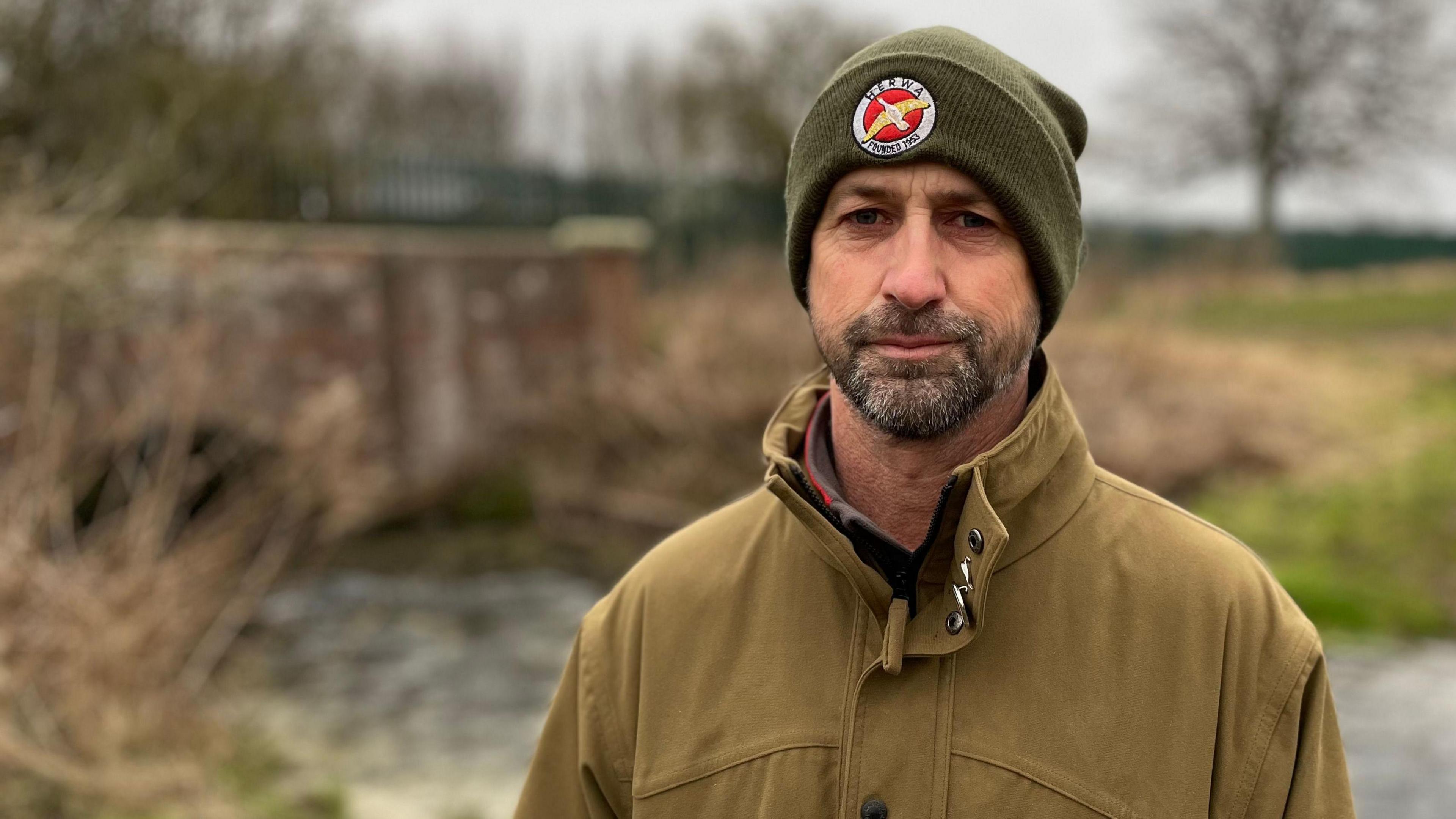 Matt Arnold frowns as he looks towards the camera. He has a dark, short beard. He is wearing a green woollen hat with a red and yellow badge on the front and a khaki-coloured coat. The backdrop is out of focus, but a stream, a red-brick bridge and trees can be seen.