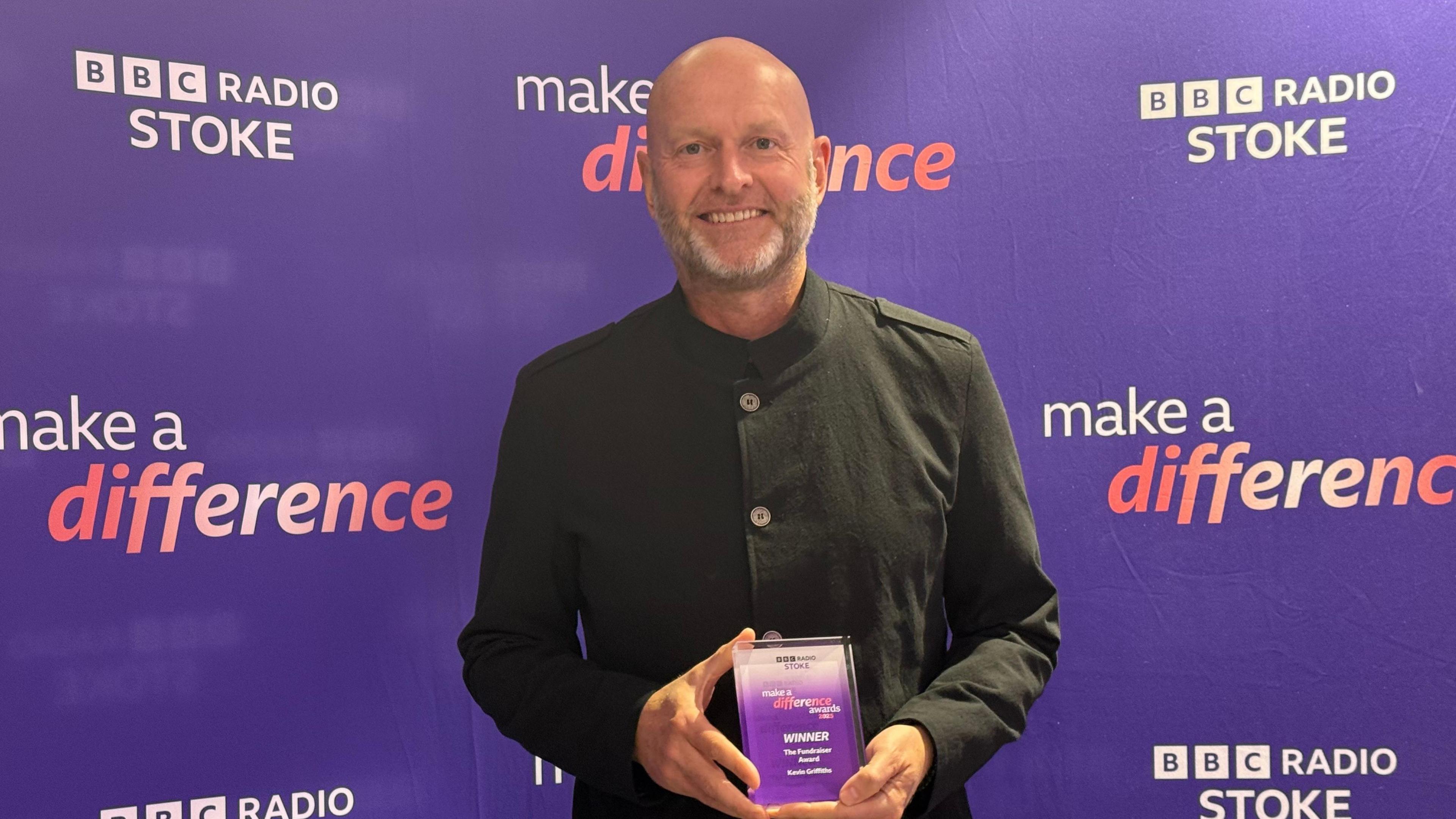 Kevin Griffiths, a man wearing a black shirt, is standing in front of a purple BBC Radio Stoke Make A Difference step and repeat board. He is holding a purple trophy and smiling for the camera.