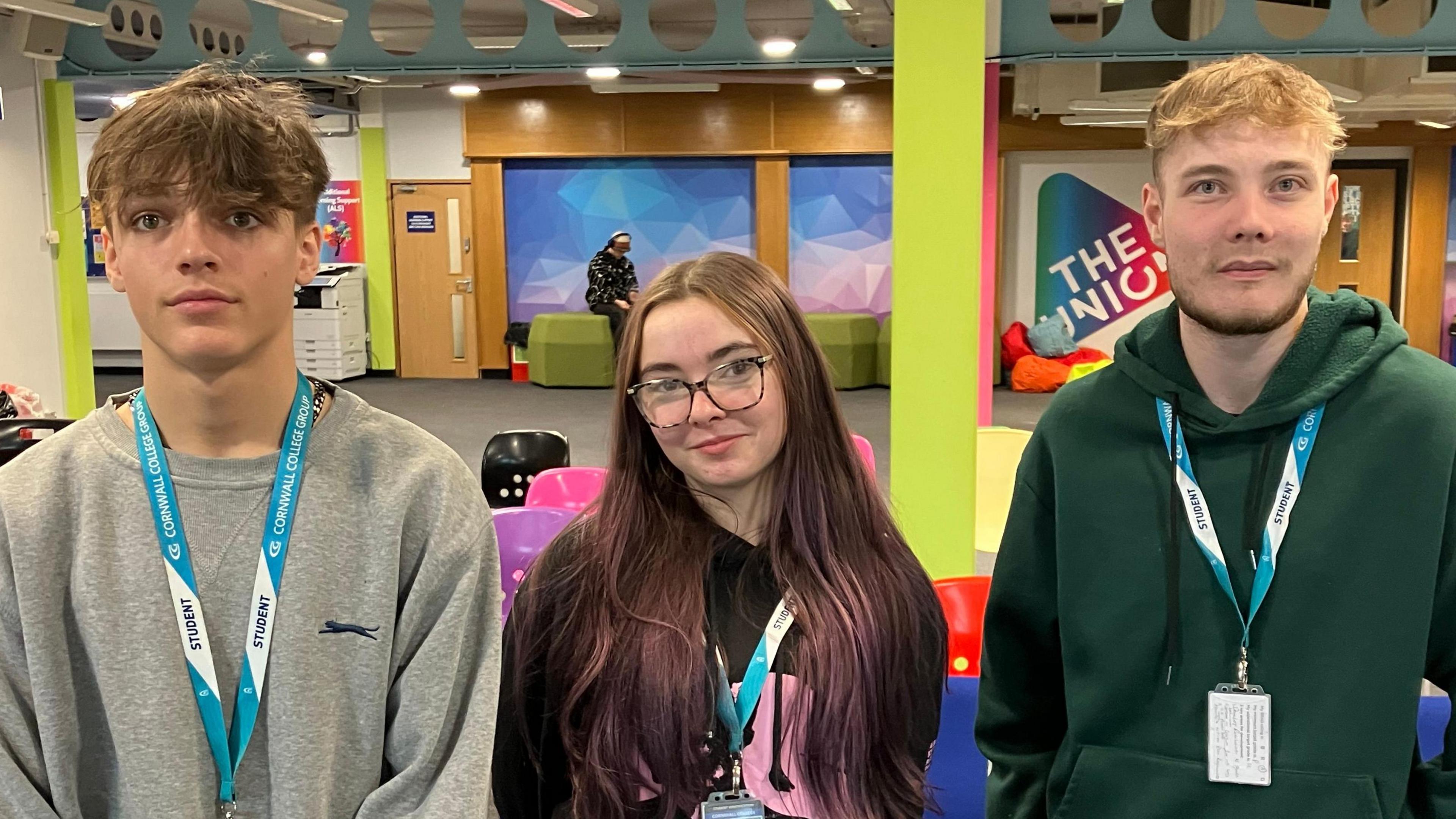 A boy with brown hair and a grey jumper on the left, a girl with dark hair and glasses in a black jumper in the middle, and a blond haired boy in a dark green hoodie on the right. They are are standing in the brightly decorated college foyer.