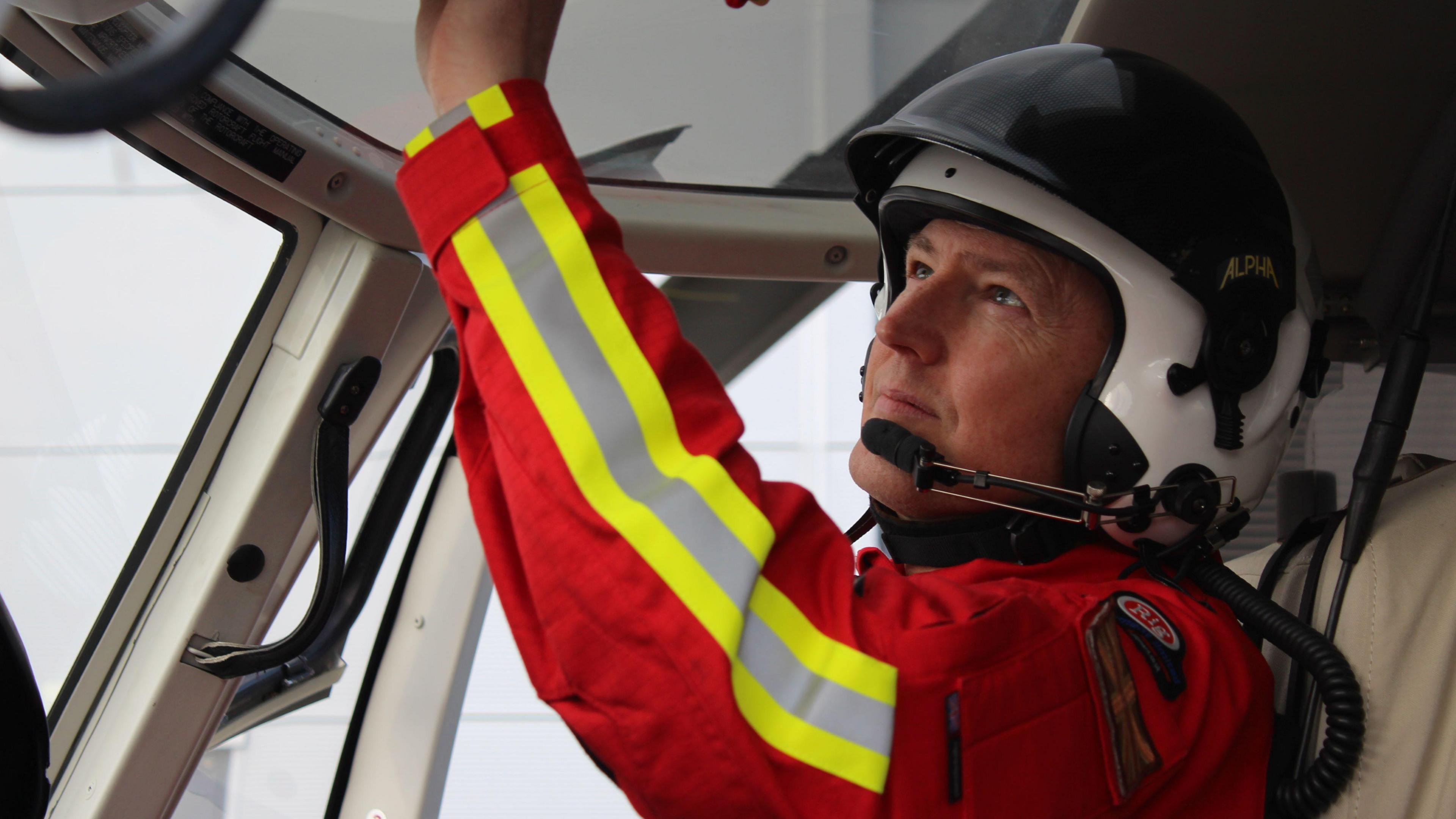 Pilot JJ Smith hits controls inside the helicopter. He is wearing red emergency service overalls and a helmet.