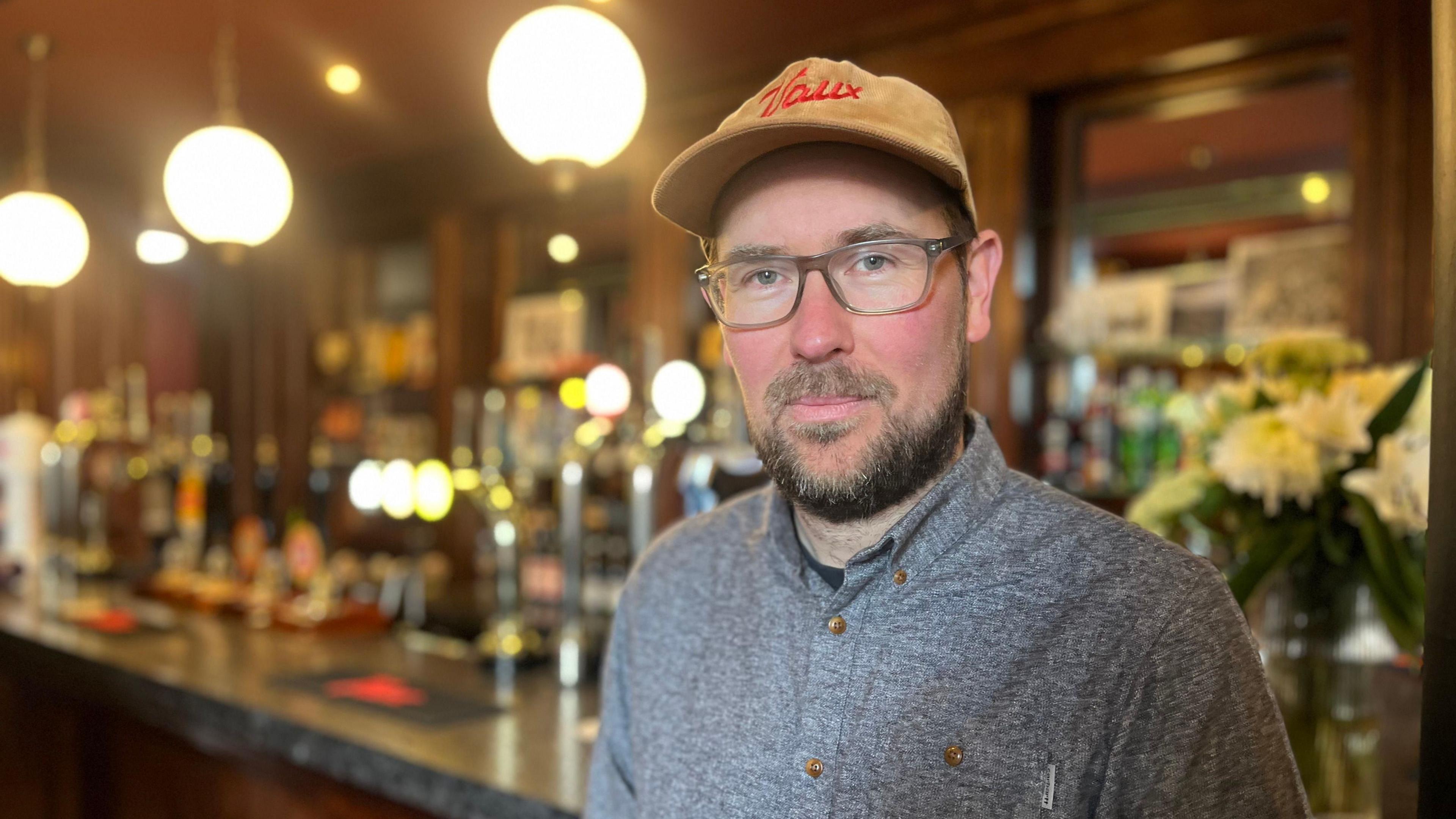 Steve Smith, wearing a cap with Vaux written on the front and a grey shirt, stands in front of the bar at the Bridge Hotel.