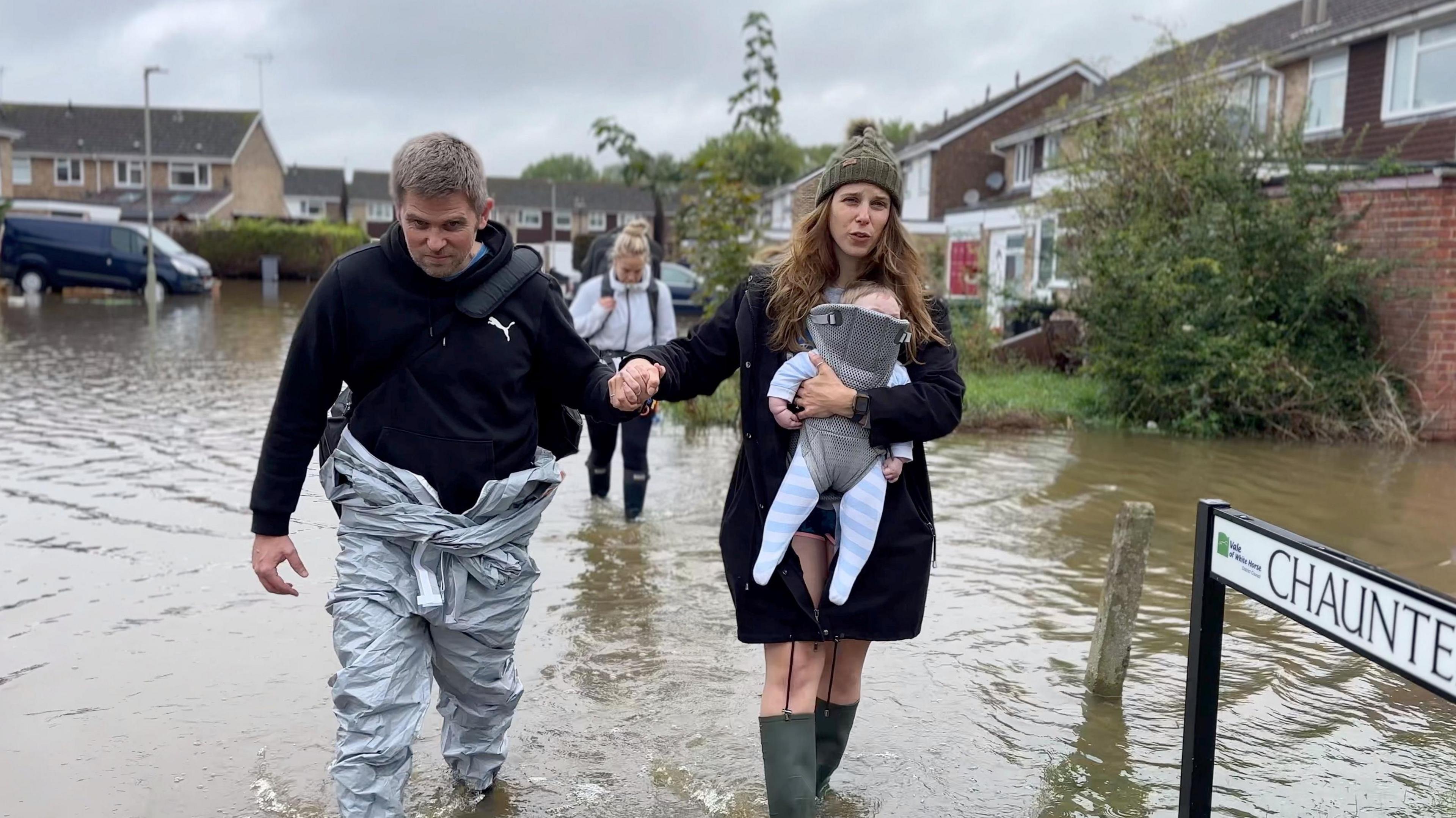 A family of three walks through flooded water in their street, Chaunterell Way, there are homes in the background and a black van to the left of the frame, the dad is standing to the left, he has short hair and wears a black hoodie with a Puma logo on the right of his chest and a silver waterproof boiler suit. He is holding a woman's hand, she wears a khaki green woollen beanie hat with a khaki green fur bobble on top of it and a black parka coat and green wellie boots, she carries a baby in a baby carrier