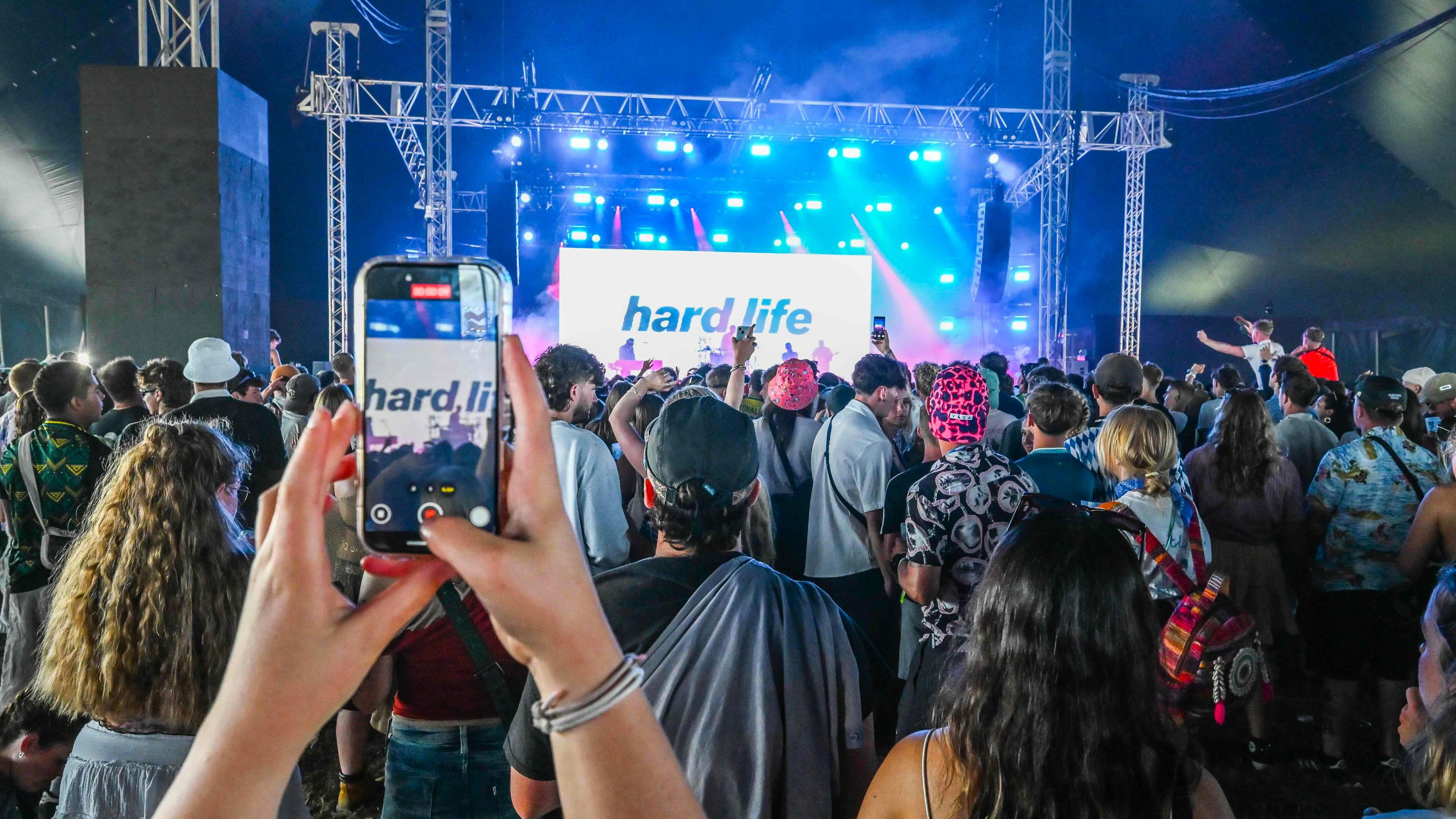 The image shows a crowd at a concert or music festival inside a large tent or covered area. The stage is brightly lit with blue and purple lights, featuring a large screen displaying the words "hard life." In the foreground, someone is holding up a smartphone, capturing the scene.
