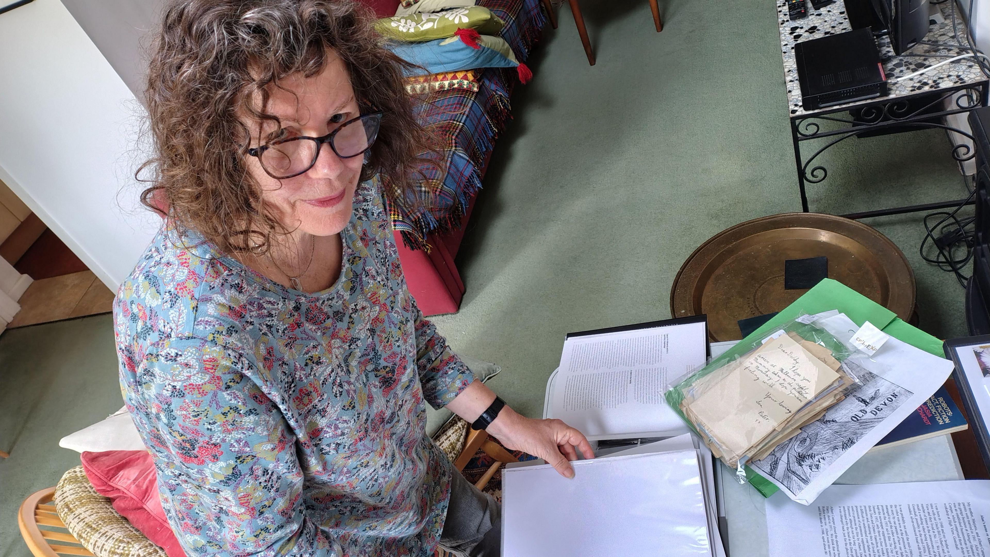A woman in a floral top with curly brown hair and glasses. She has a stack of papers in front of her.