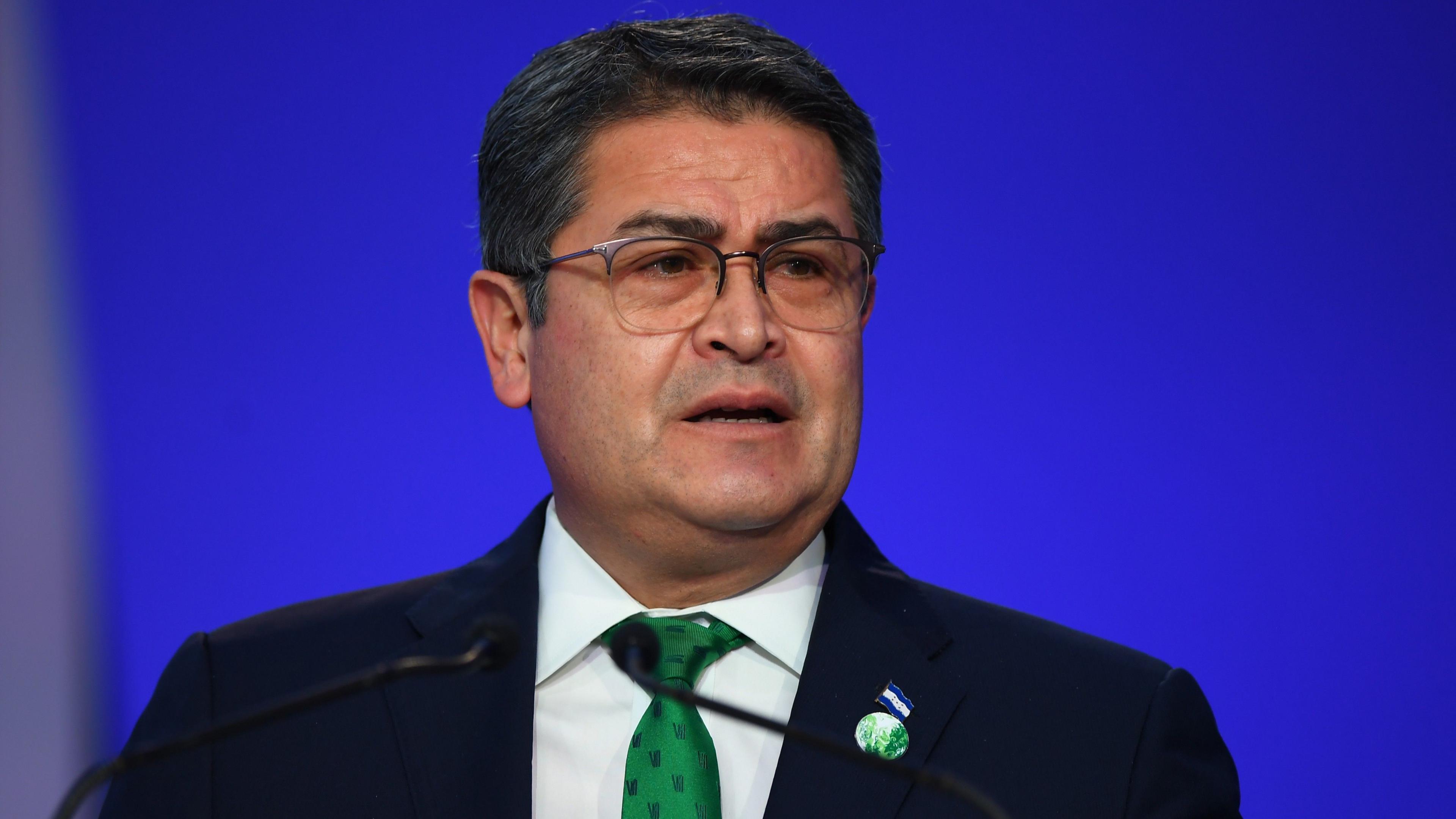 Honduras President Juan Orlando Hernández speaks in front of a blue backdrop. He has short hair and wears square-framed eye glasses. 
