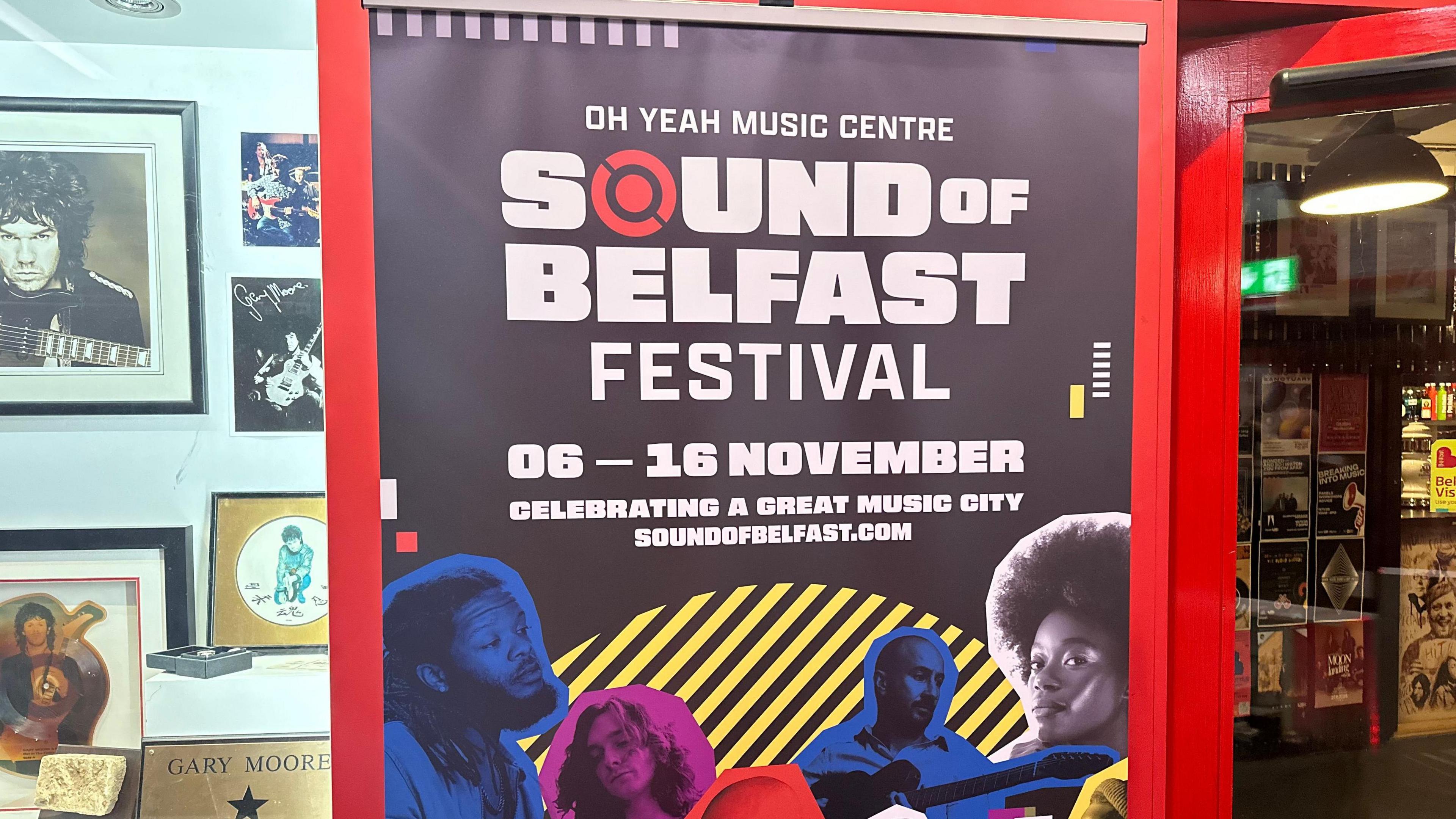 Poster with Sounds of Belfast written on it. It is against a all with pictures and beside a glass door 