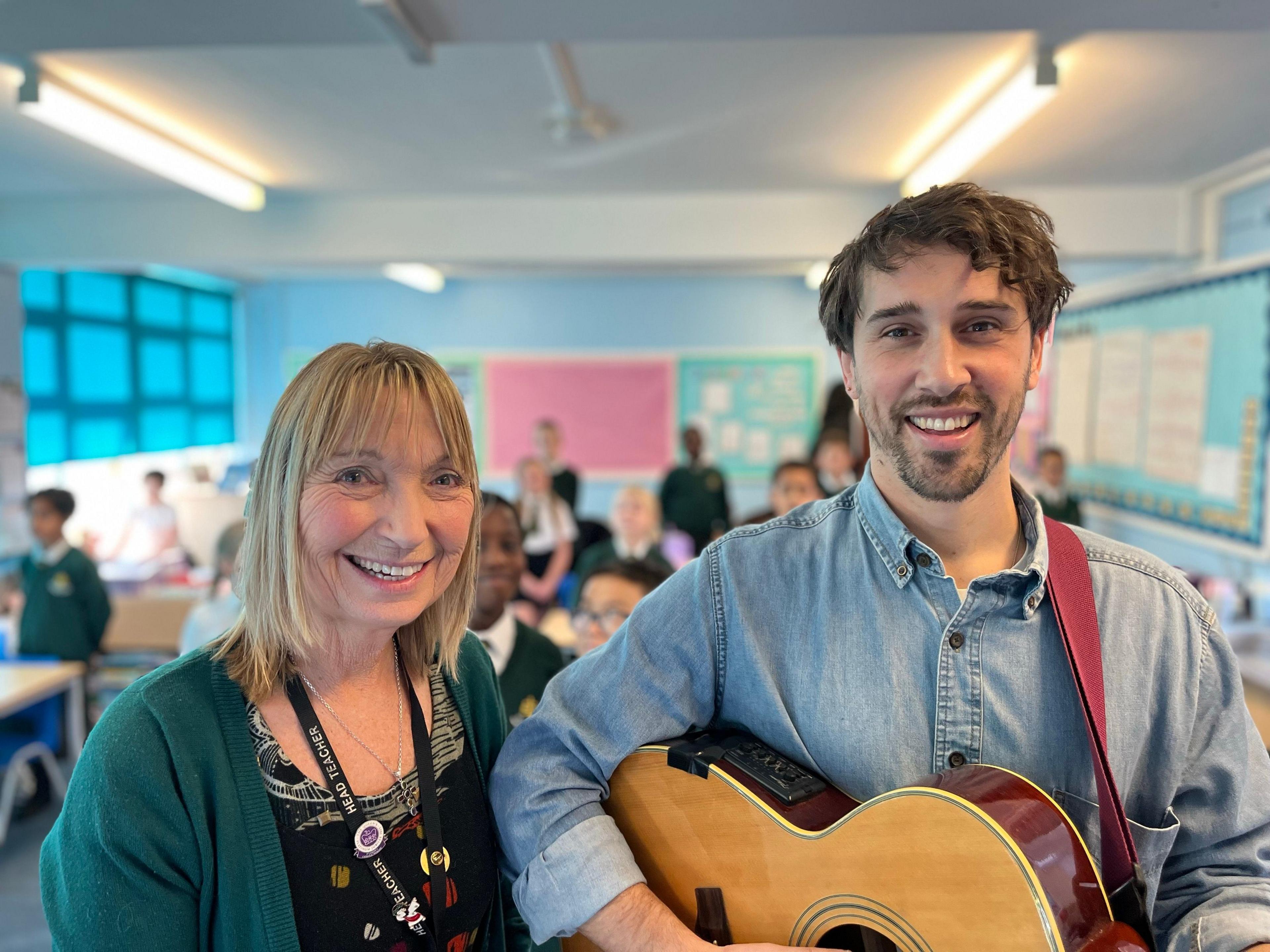 Two teachers standing in the foreground with one of them holding an acoustic guitar with children behind in the background across the classroom