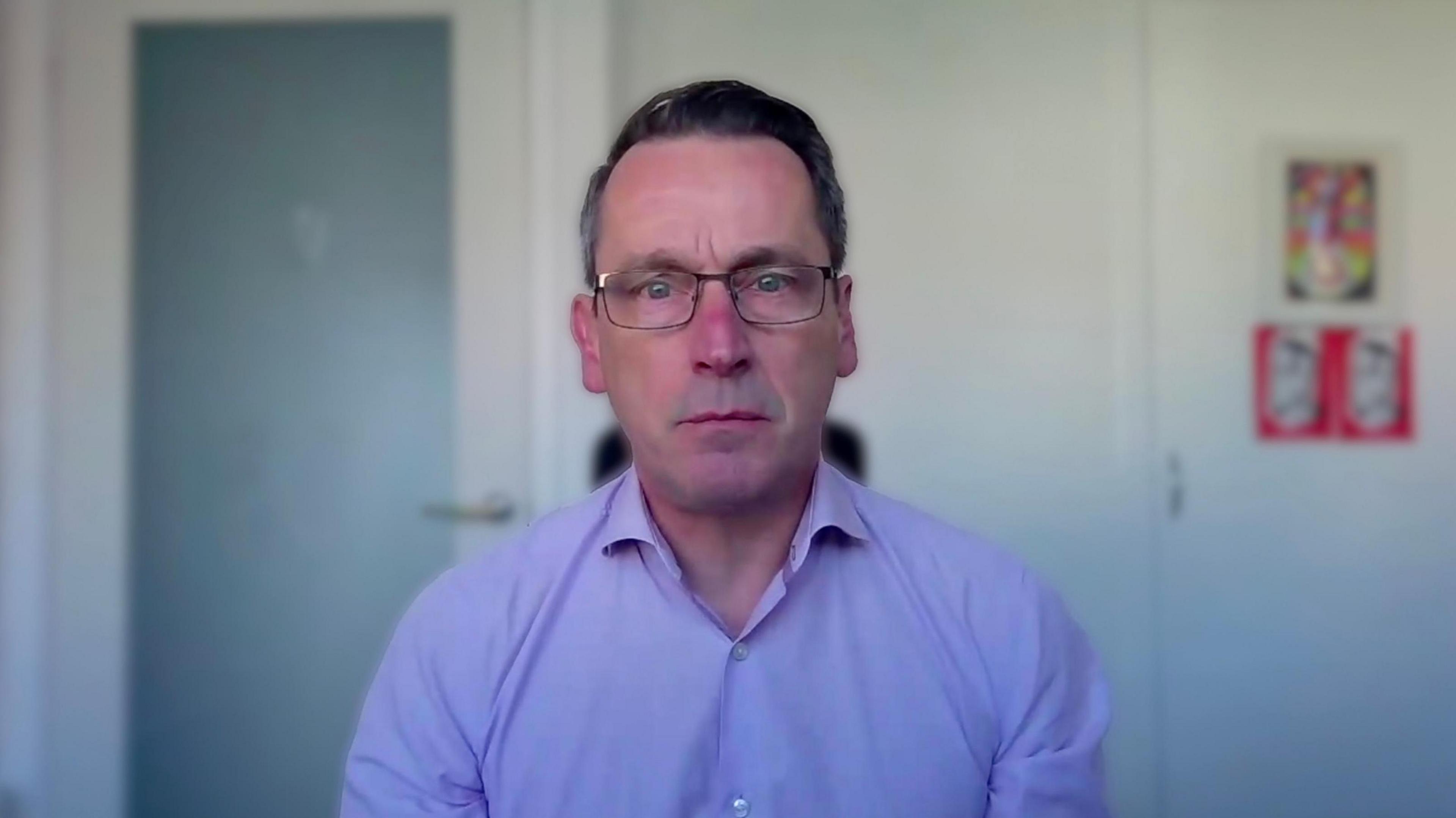 A man with glasses and a light blue shirt is sitting in his home office doing an online interview 