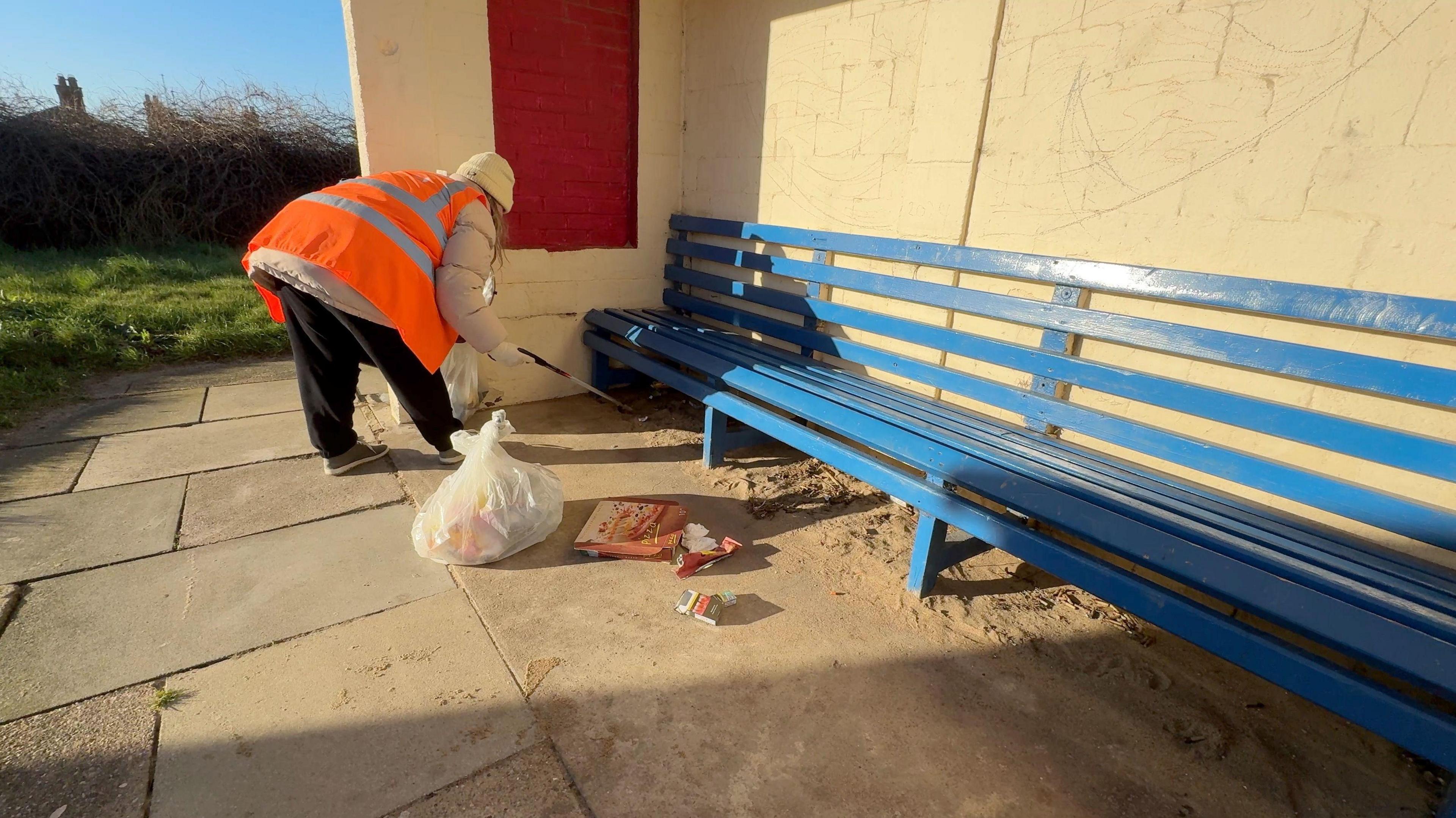 A woman with a litter picker reaching underneath a blue bench. She is wearing black trousers, an orange high vis jacket and beige beanie. Next to her is a white full plastic bag and a pizza box, cigarette packet and plastic next to her.