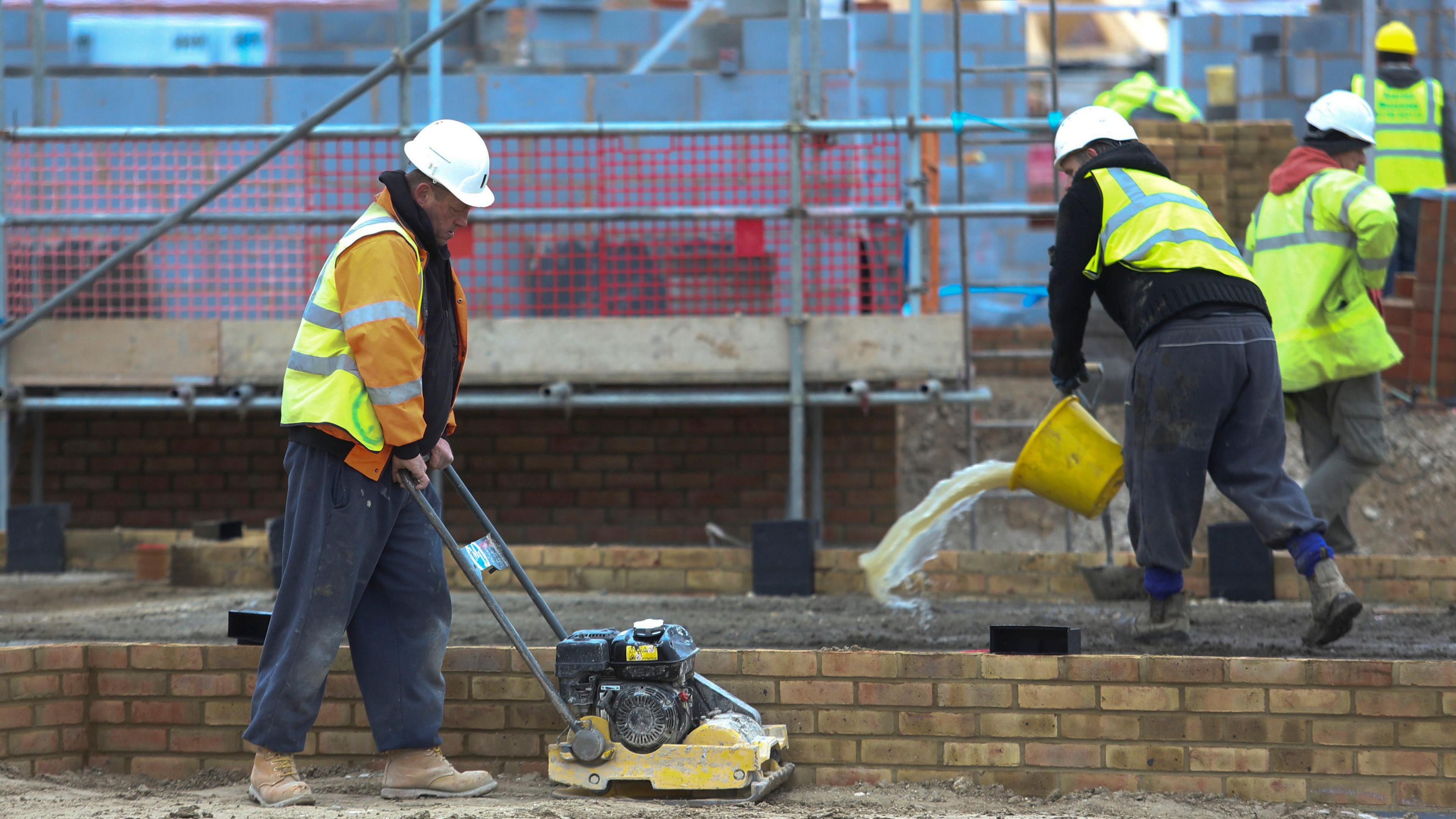 Four male construction workers on a building site. All are wearing hard hats and hi-vis jackets, and carrying out a range of tasks using different tools. 