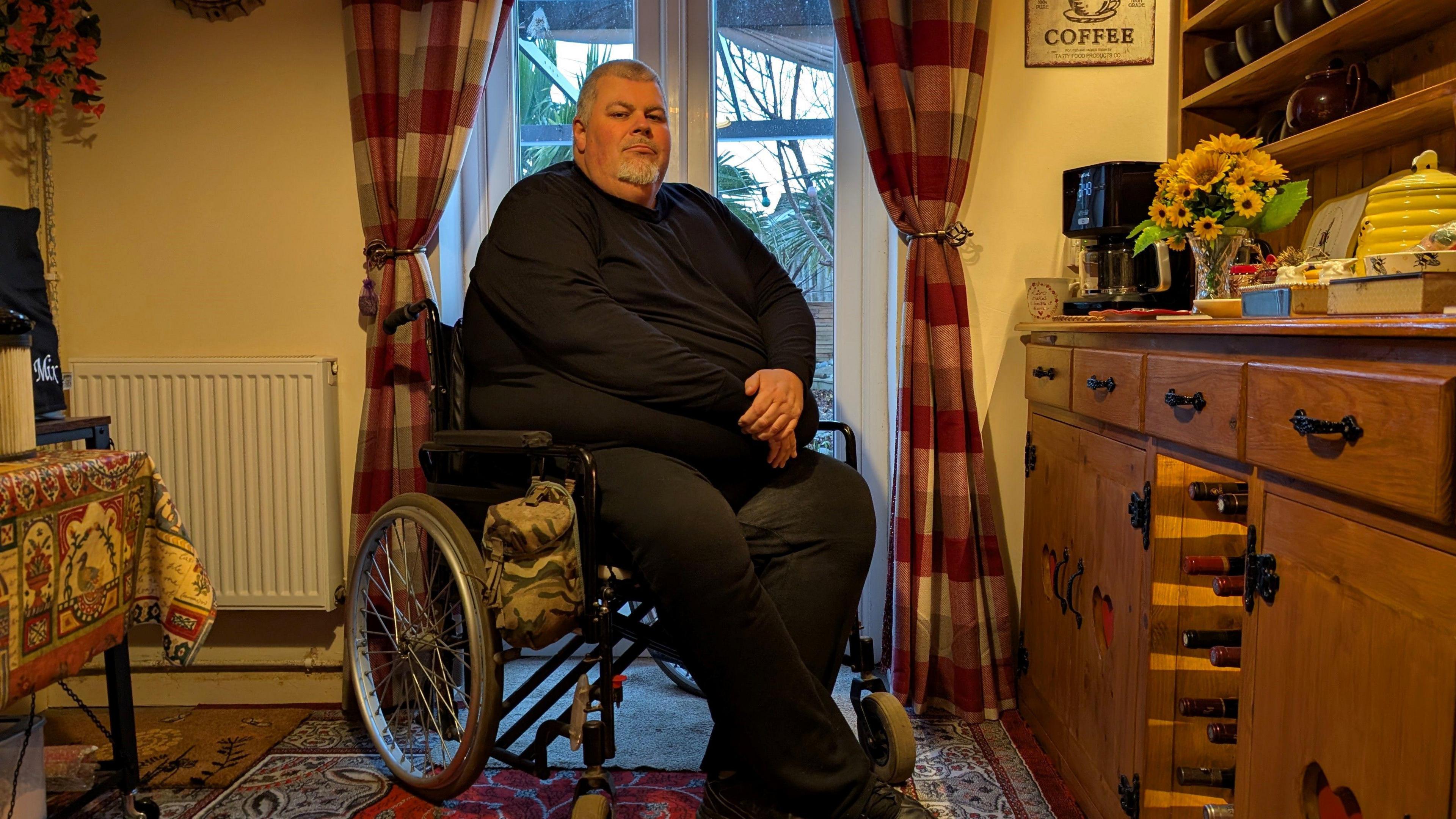 Neal Wylde, wearing all black, is sat in front of French windows in a kitchen