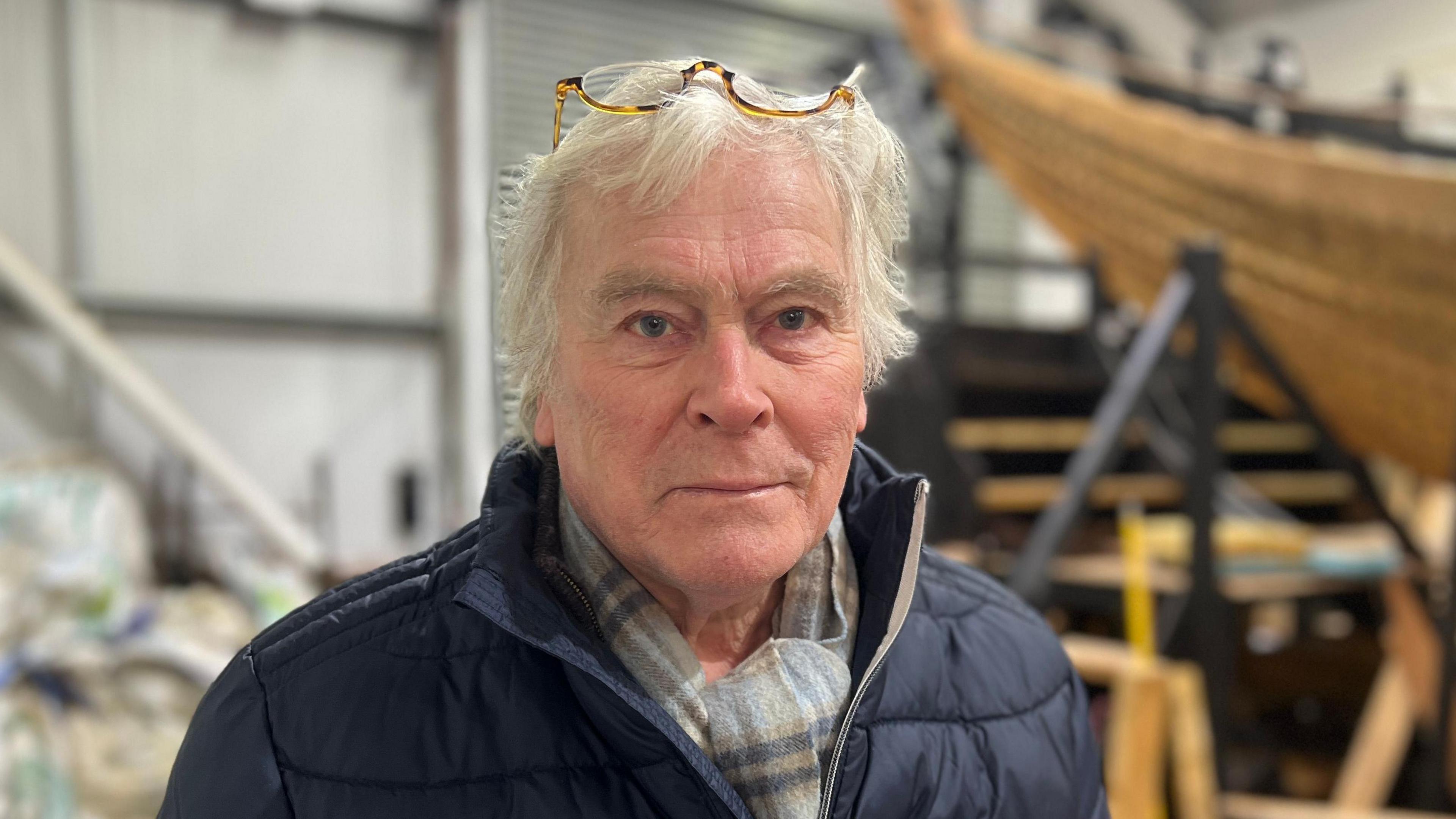 Sean McMillan smiles at the camera posing next to the longship build. He has grey hair and glasses sit on top of his head. He wears a navy coat with a chequered light blue scarf.