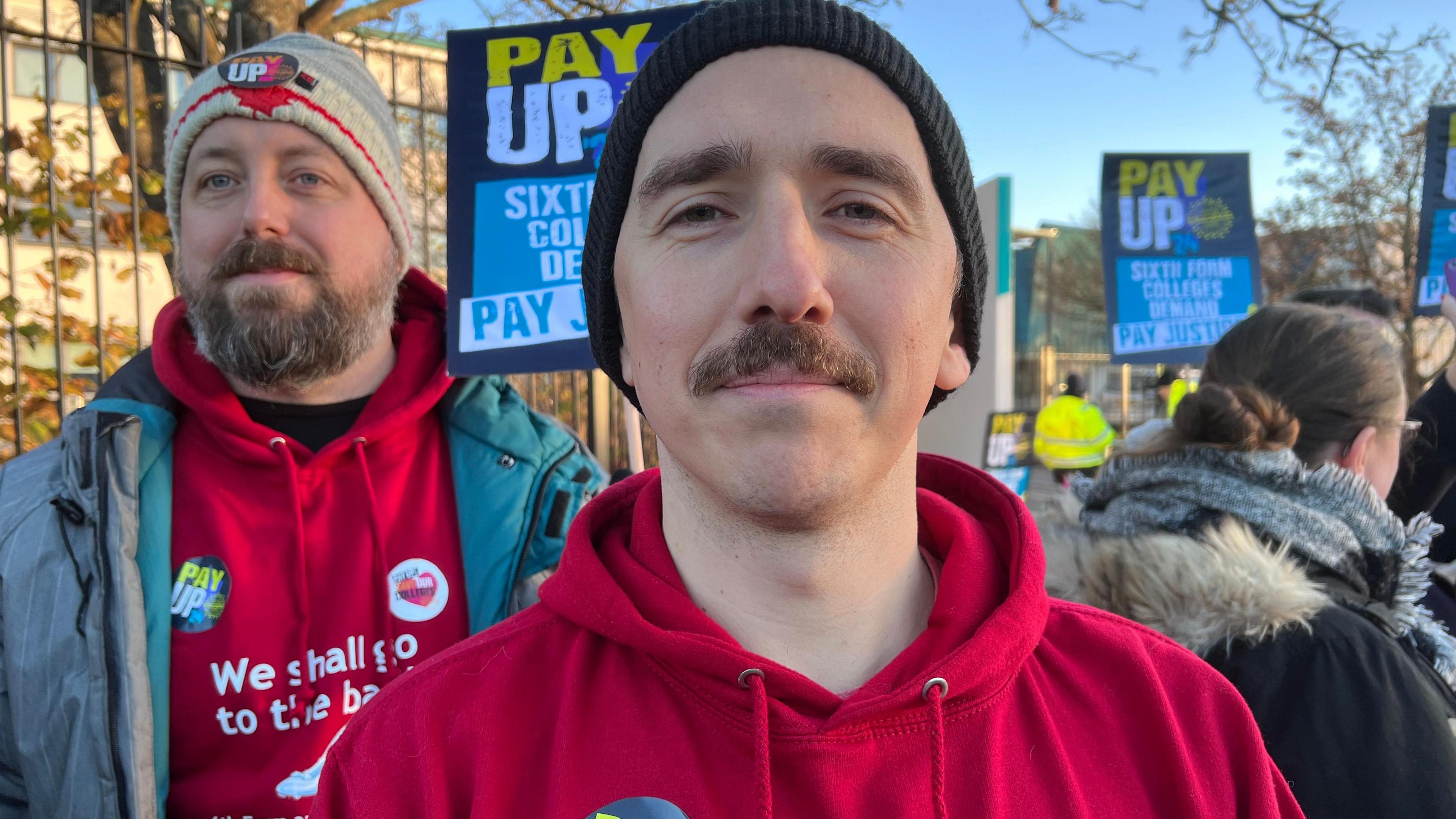 A man in a red hoody jumper standing in front of a picket line. Behind him are banners saying PAY UP