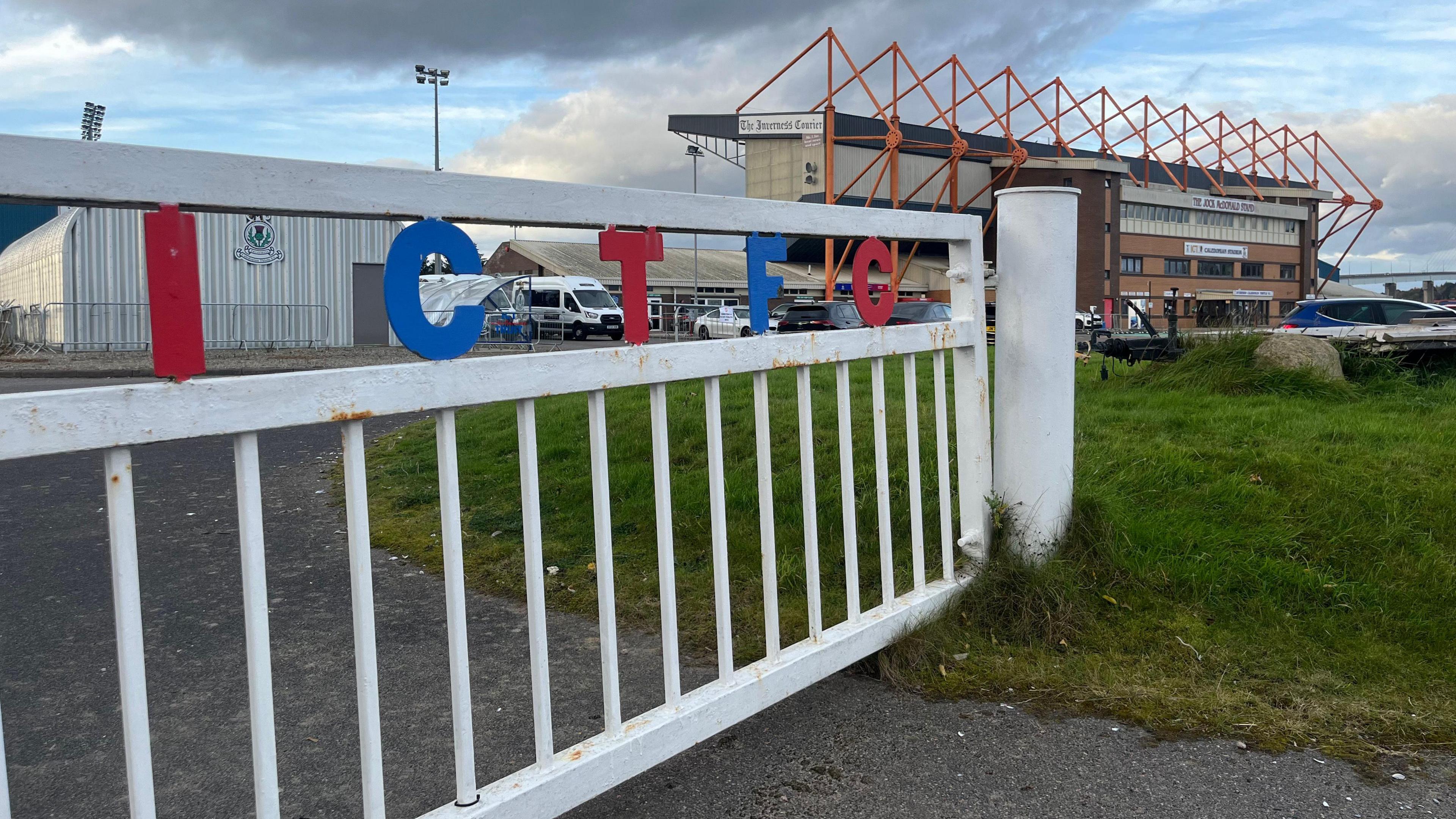 Outside the Caledonian Stadium. A white fence with the letters 'ICTFC' in alternating red and blue colours is on top. In the background, the stadium can be seen.