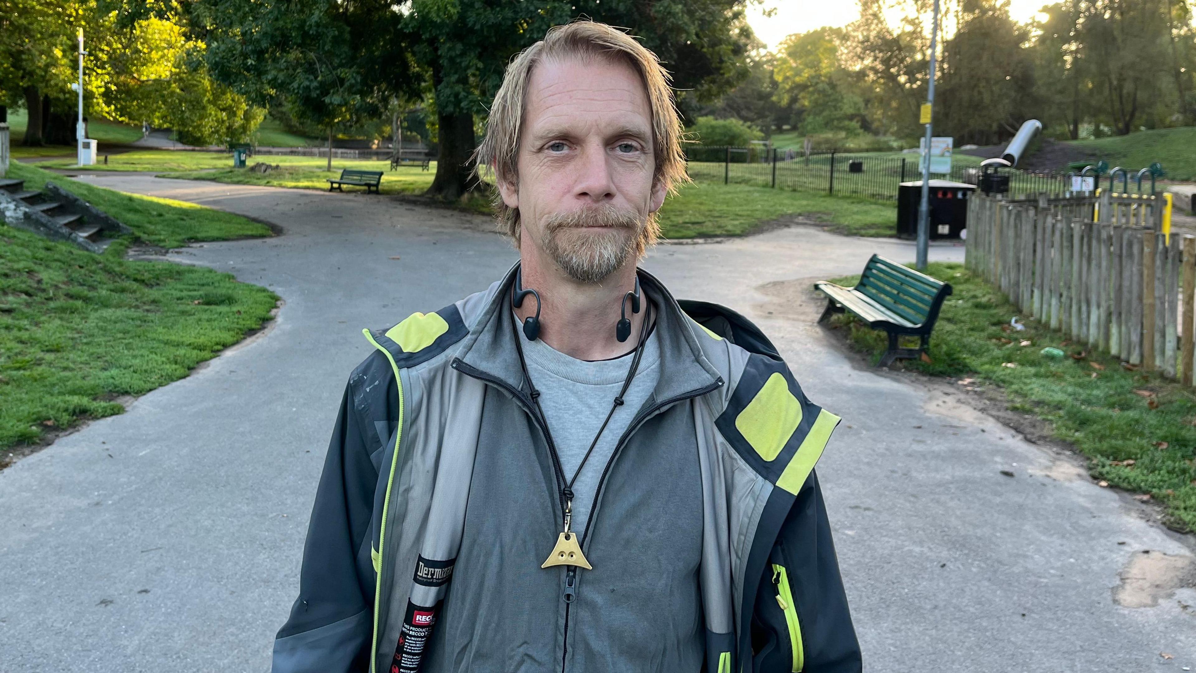 Matt Peters looking at the camera with a goatee, several layers on clothing including a rain coat and hoody, and some earphones. He is stood on a pathway in St George Park next to the play park.