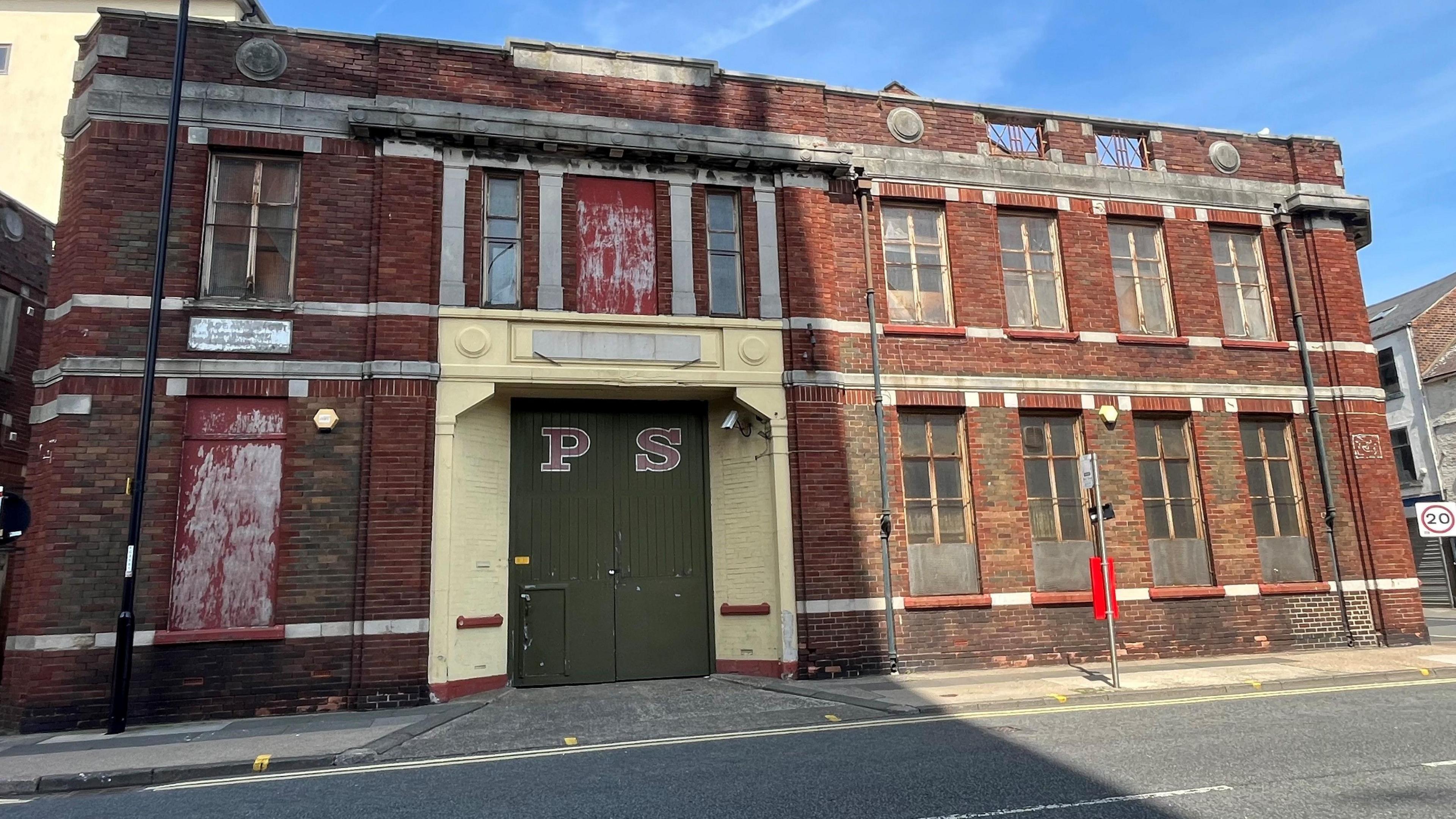 The currently derelict building of the proposed Glassworks site. It is a two-storey, red brick, old factory building with PS written on the green doors.